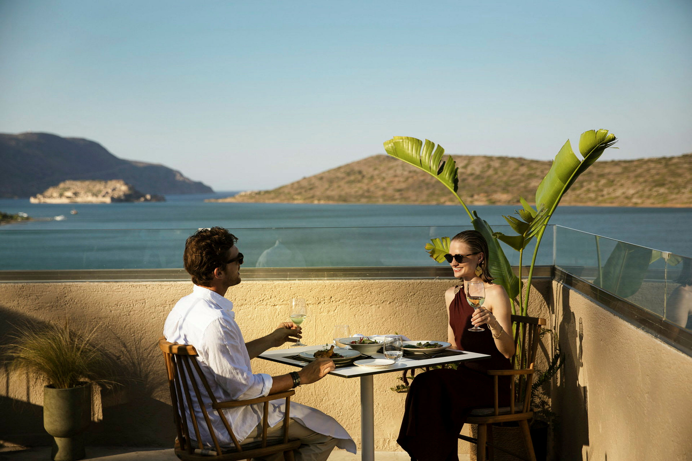 To personer sidder ved et bord med mad og drikke på en terrasse med udsigt over havet og bjergene. (AI-genereret tekst)