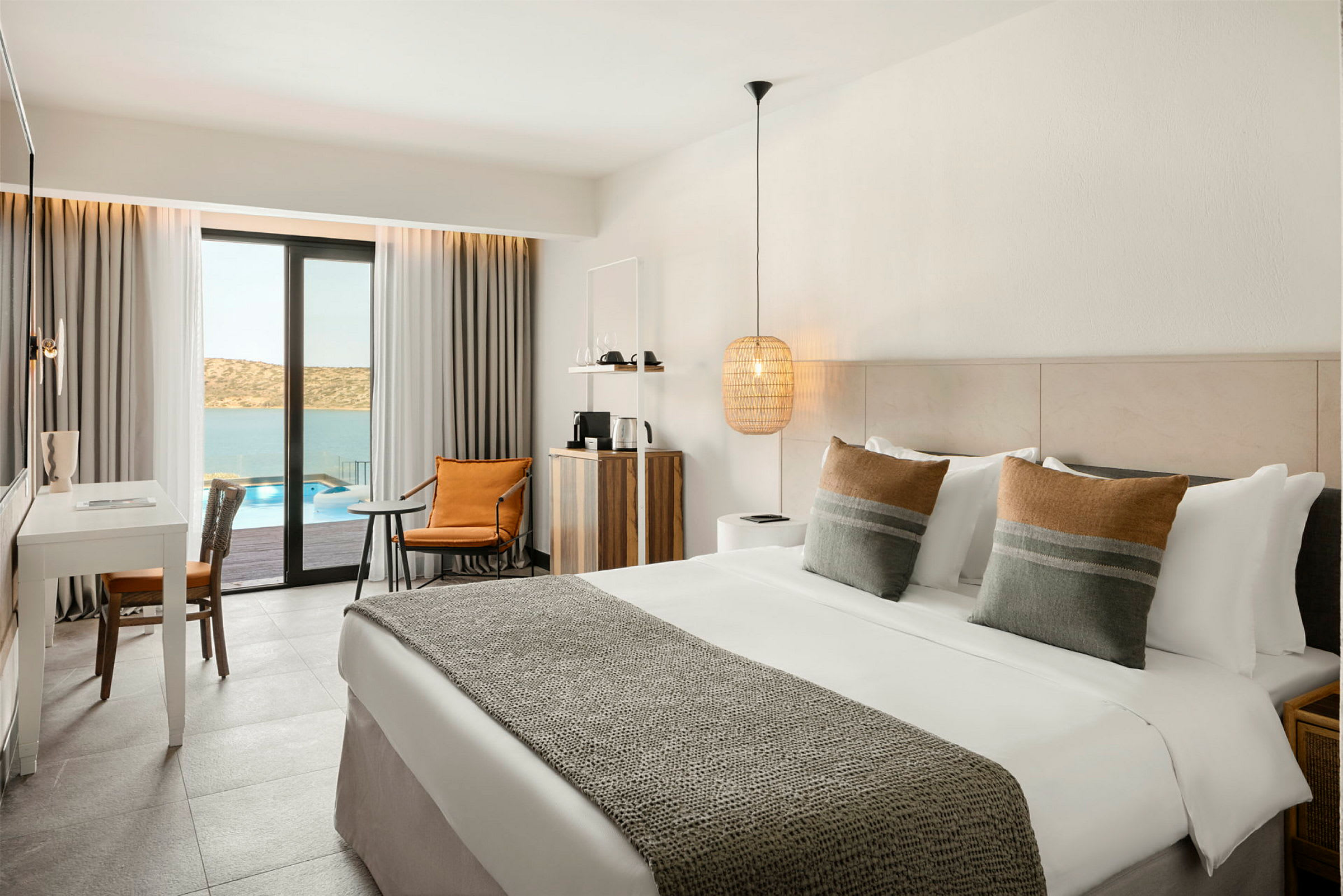 Hotelværelse med dobbeltseng, skrivebord og siddeområde, med udsigt over pool og hav. (AI-genereret tekst)