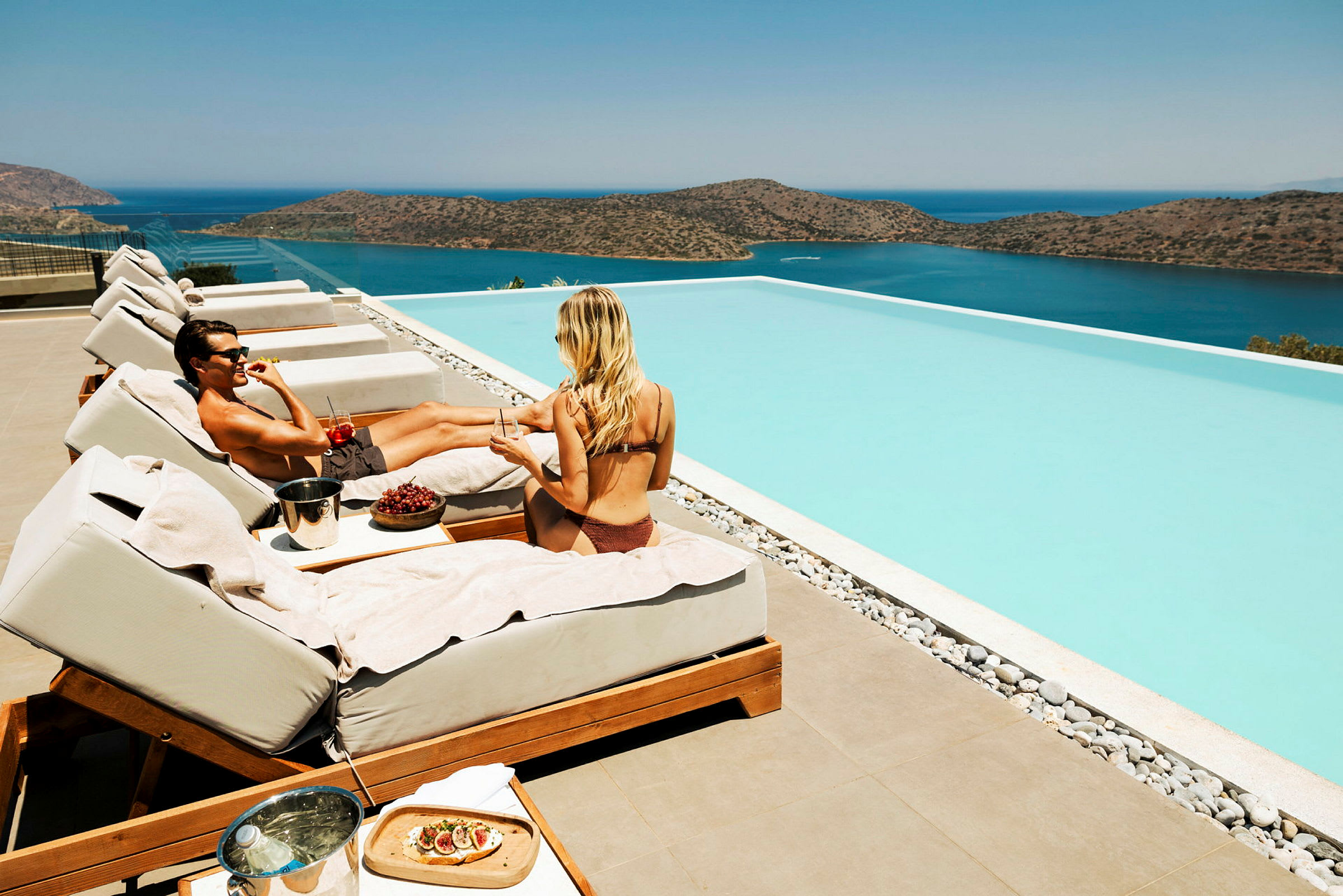 To personer sidder på solsenge ved en infinitypool med udsigt over en sø. (AI-genereret tekst)