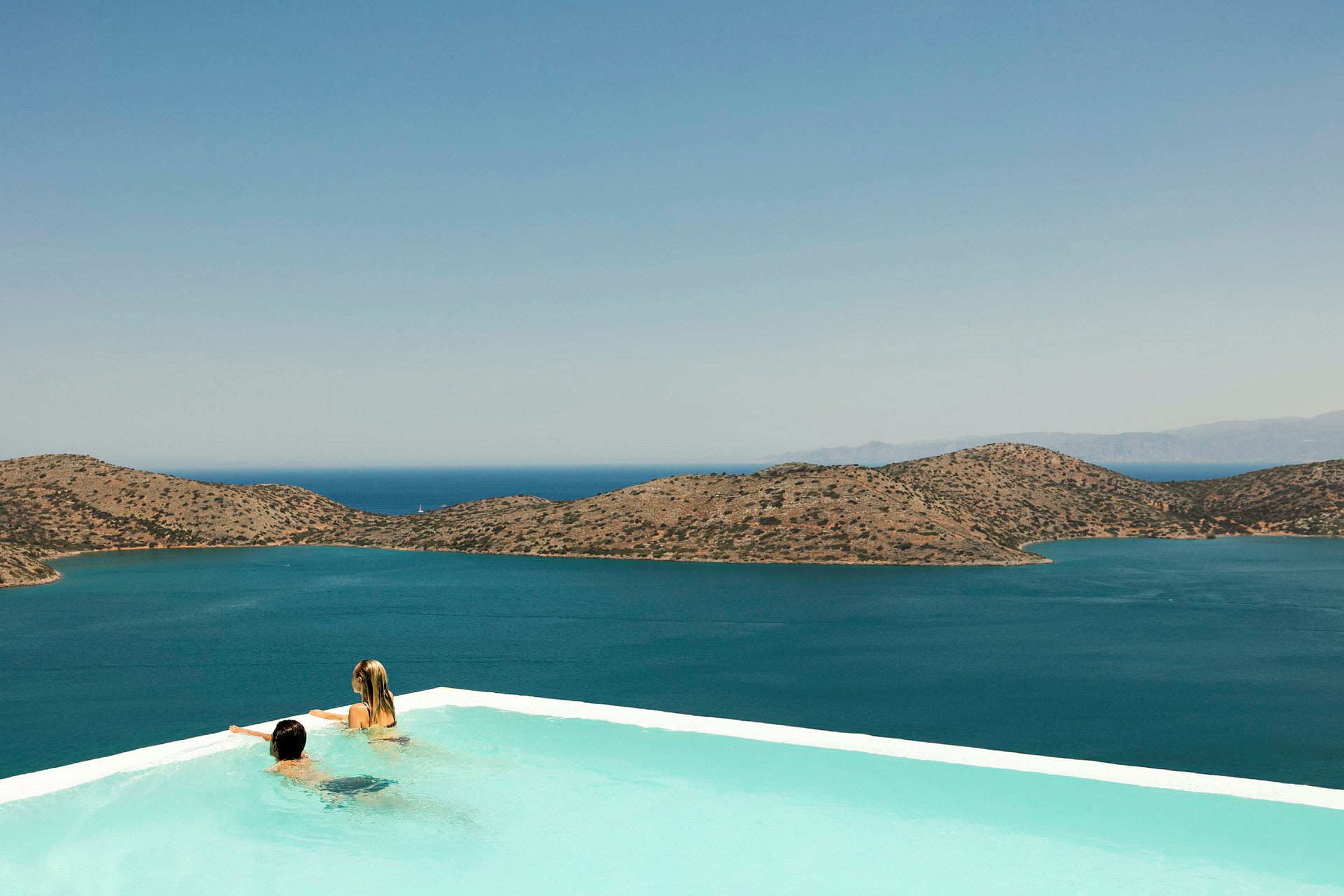 To personer i en infinitypool med udsigt over havet og bjerge i baggrunden. (AI-genereret tekst)