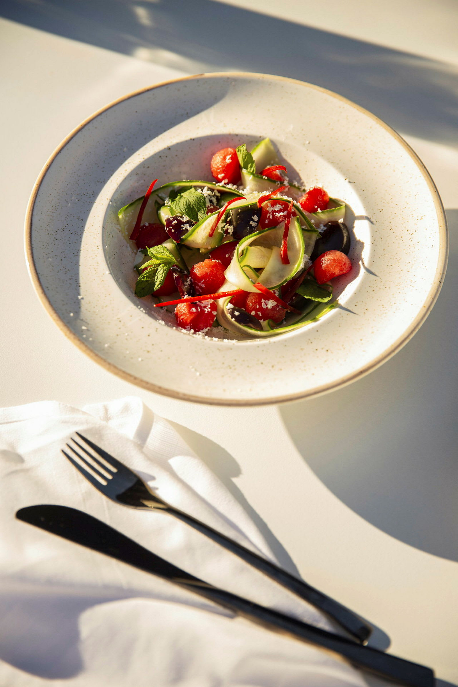 En skål med vandmelon, courgette og urter på et bord med bestik. (AI-genereret tekst)
