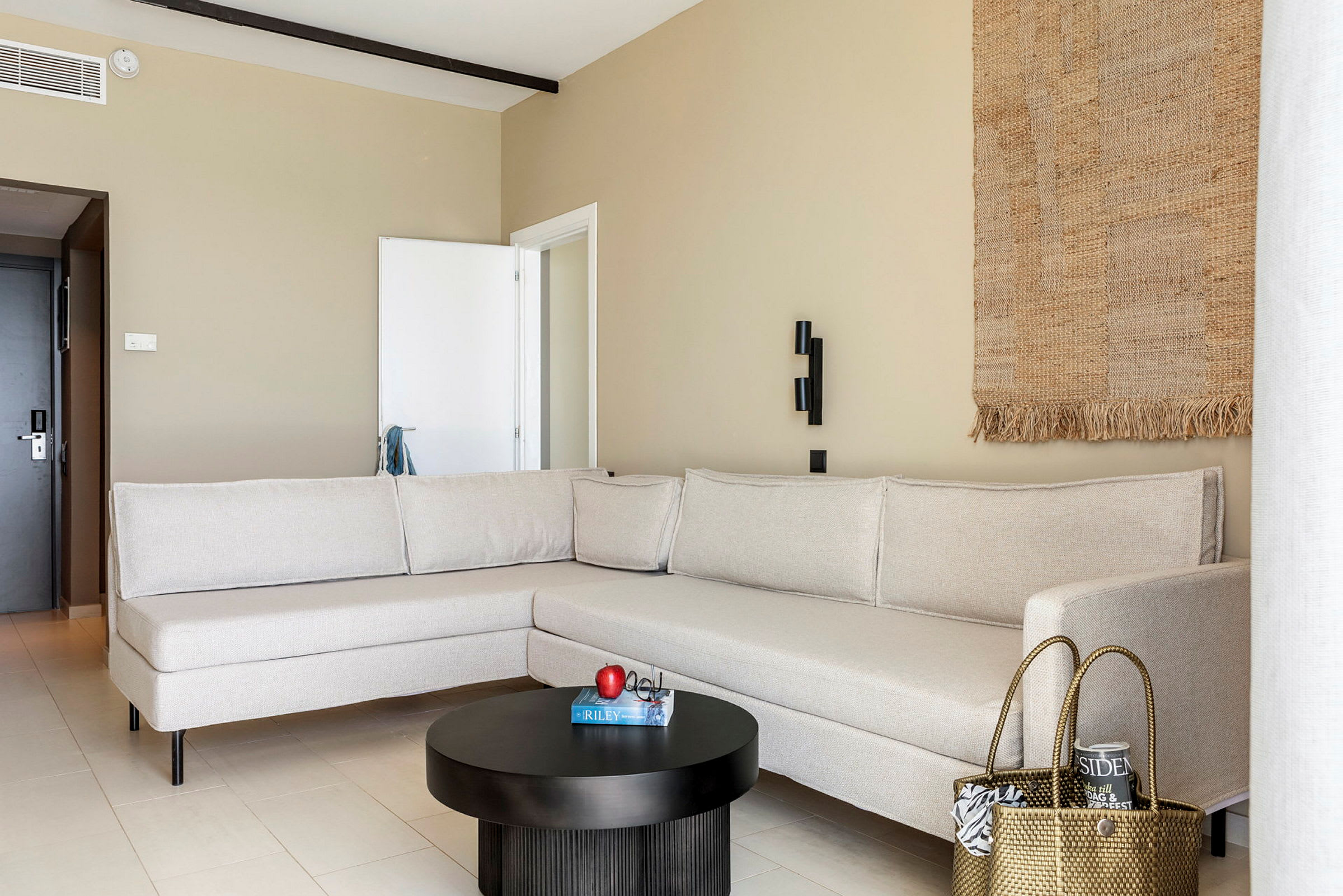 En moderne stue med en beige hjørnesofa, et sort rundt sofabord med et æble og en bog ovenpå, og et vævet dekorativt vægtæppe. (AI-genereret tekst)