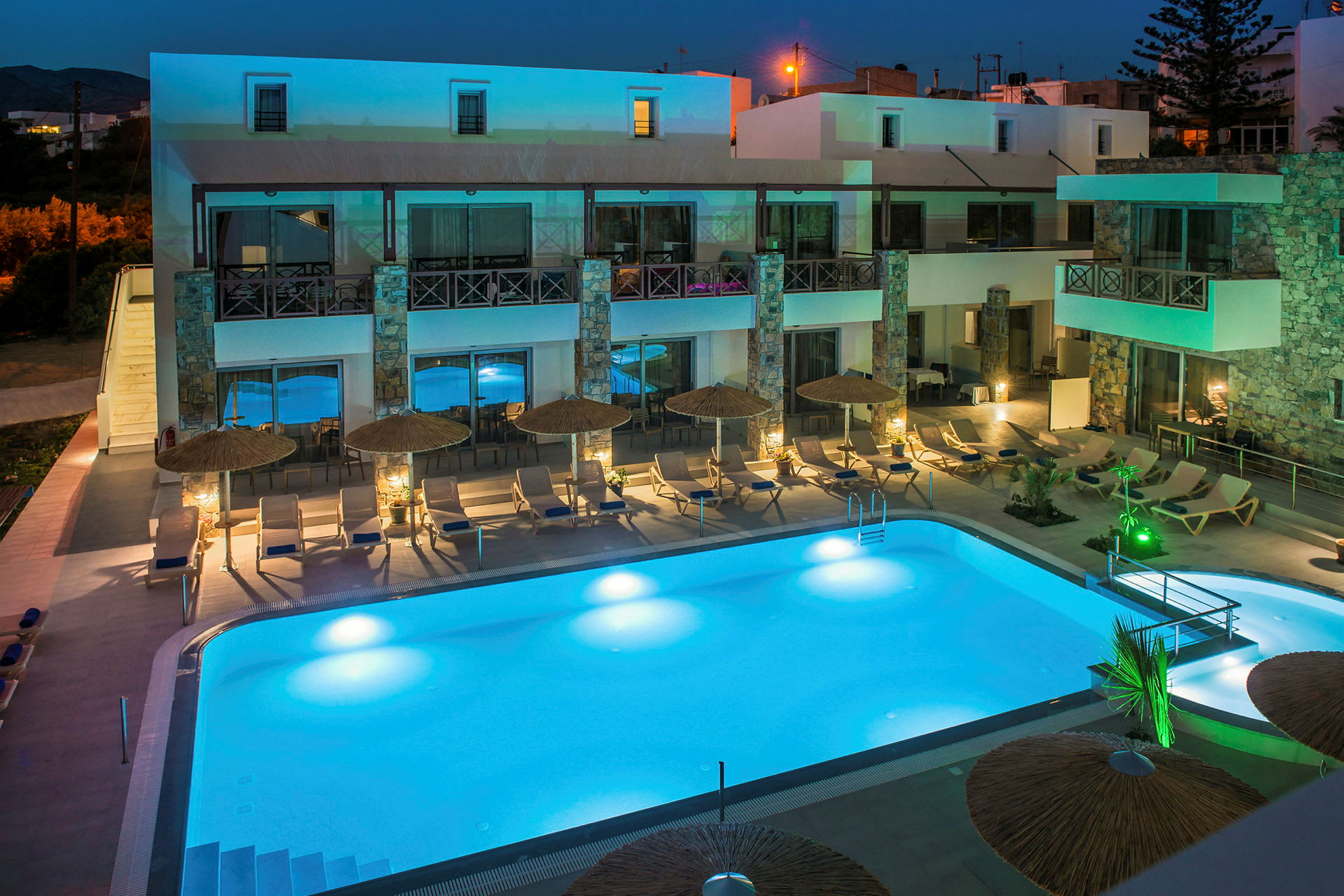Hotellet i aftenbelysning med oplyst poolområde og liggestole. (AI-genereret tekst)
