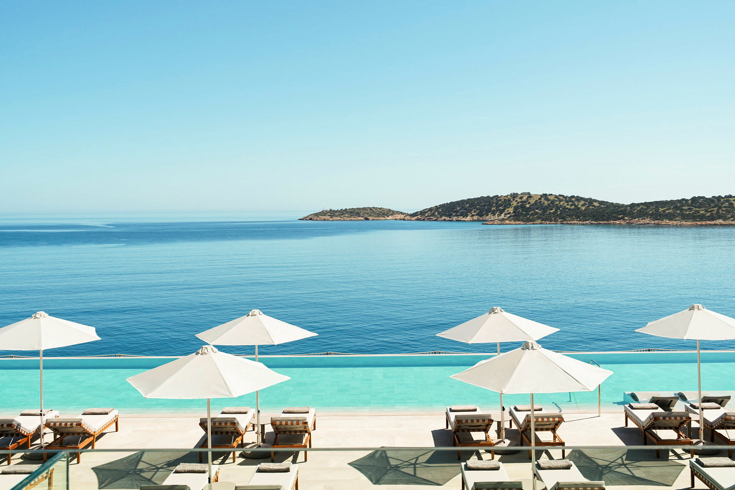 Solsenge og parasoller ved en pool med udsigt over havet og en ø i baggrunden. (AI-genereret tekst)