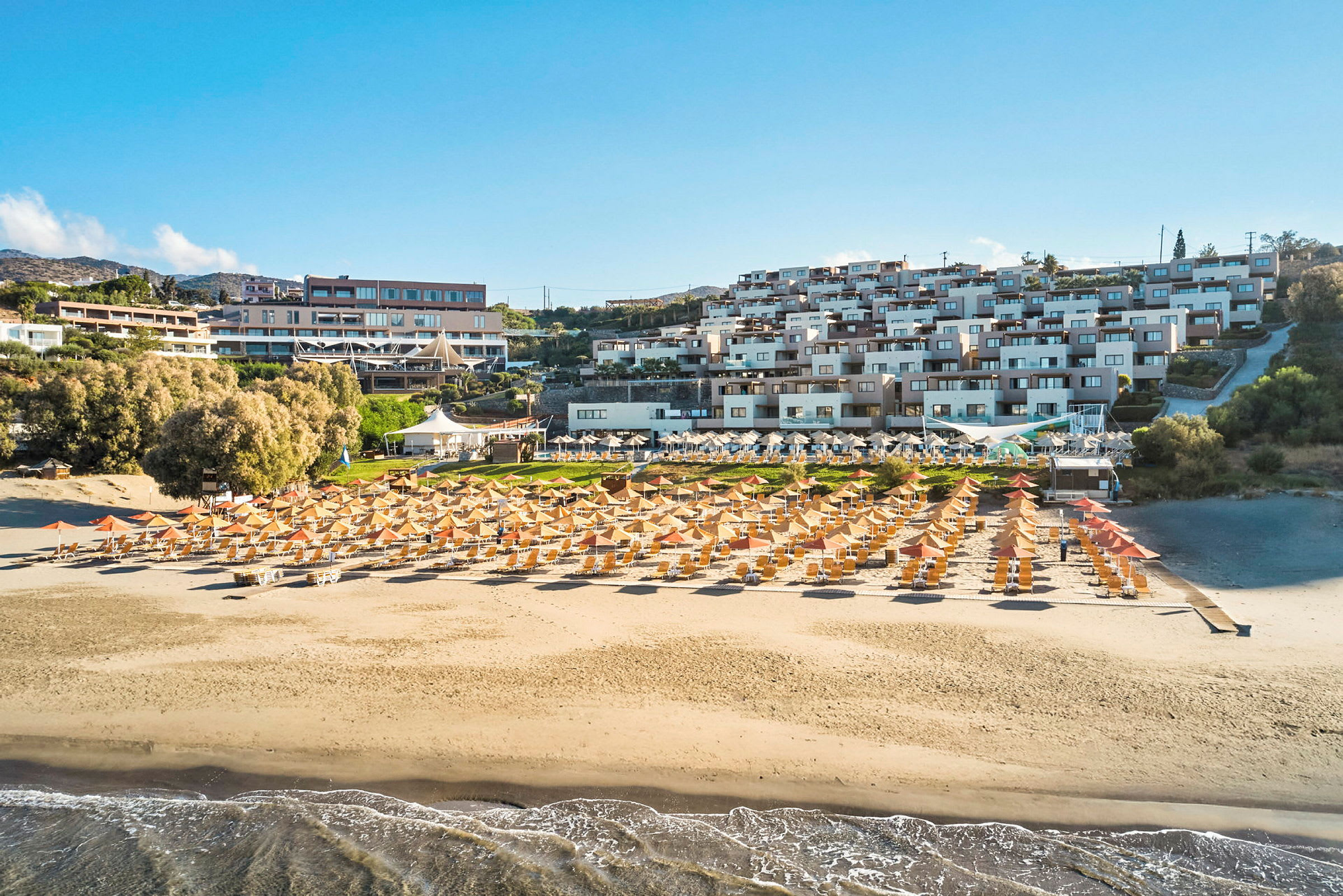 En sandstrand med liggestole og parasoller foran en resort med flere bygninger. (AI-genereret tekst)