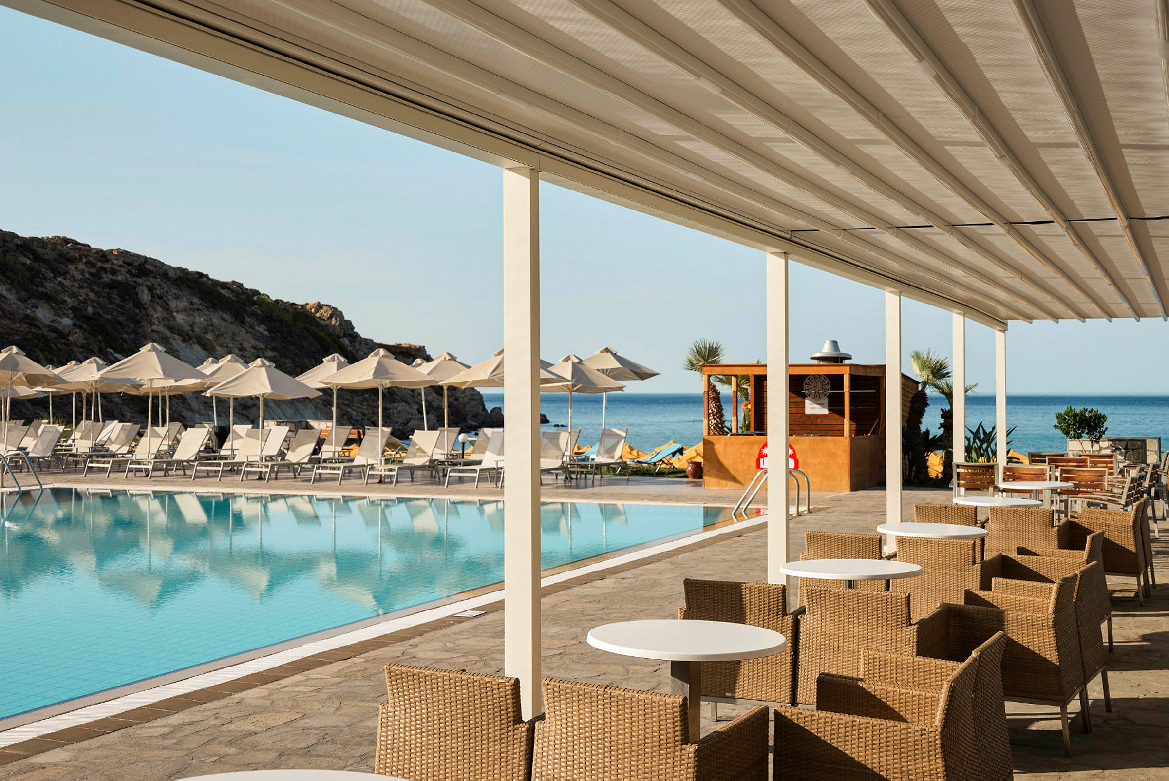 Billede af et poolområde ved en resort med solsenge og parasoller foran havet. Foran poolen er der siddepladser med borde og stole under et overdække. (AI-genereret tekst)