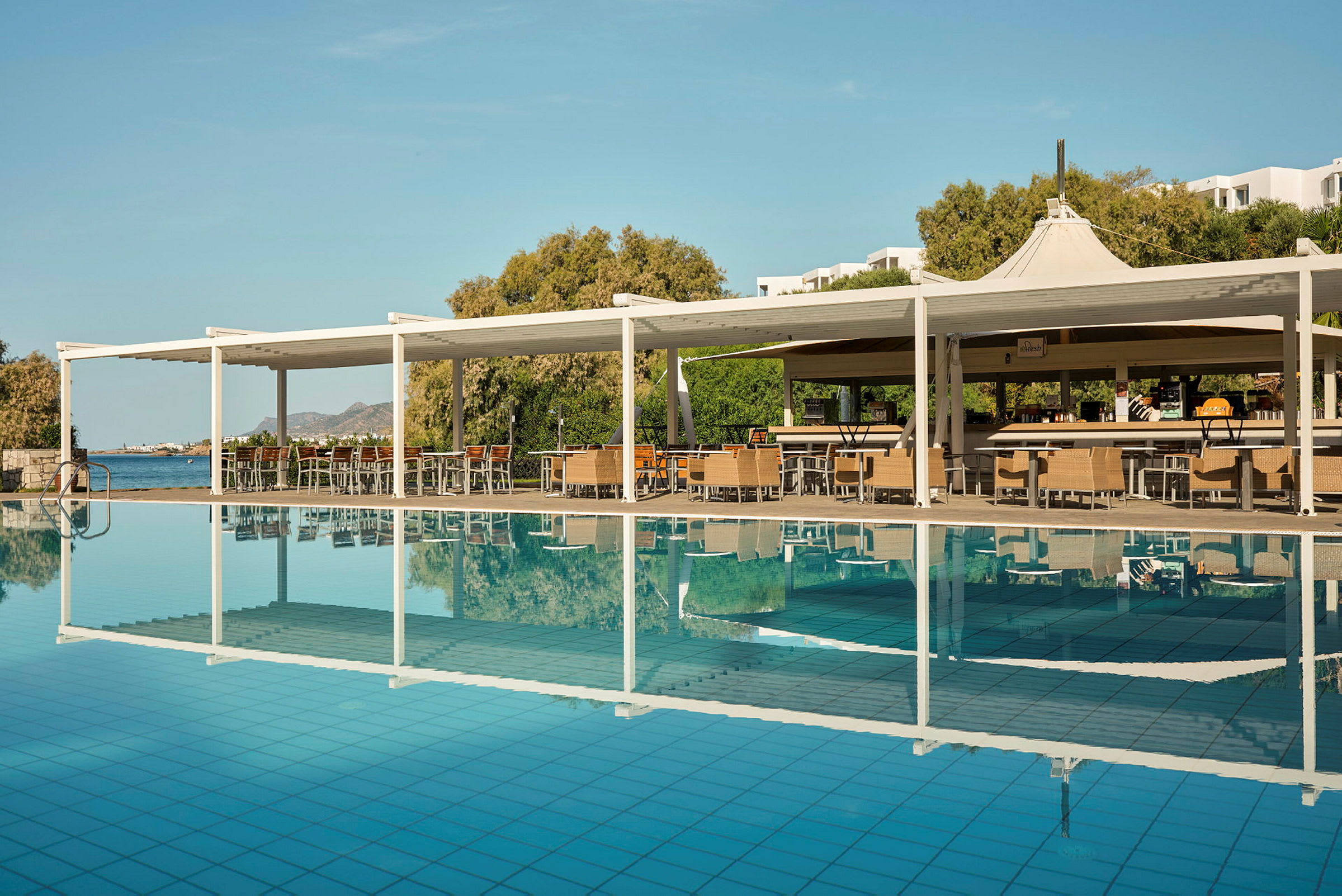 Billede af en pool med en tilhørende siddeplads og bar under en hvid pergola. I baggrunden ses frodige træer og havet. (AI-genereret tekst)