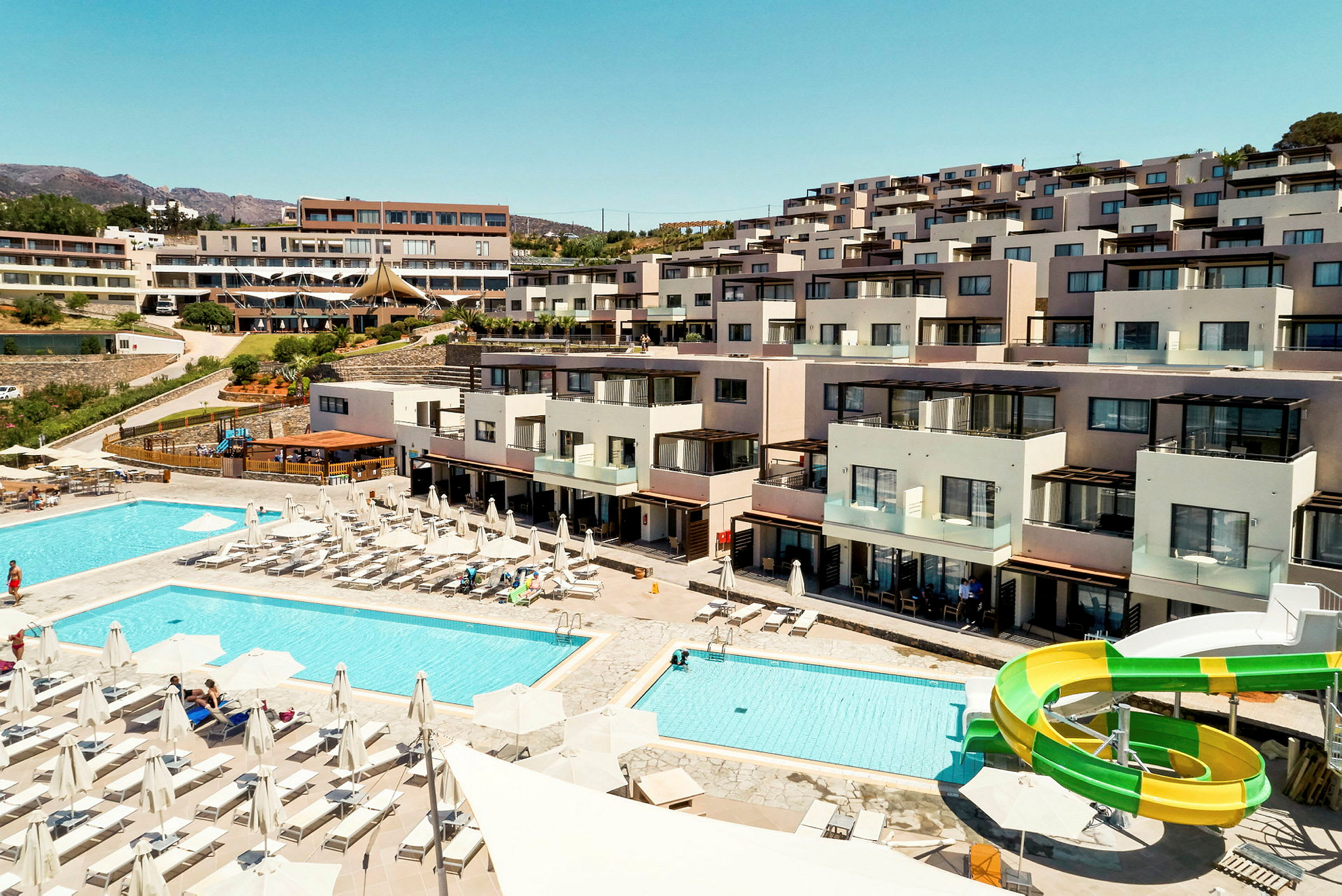 Hotelområde med pools, liggestole og vandrutsjebane foran bygninger. (AI-genereret tekst)