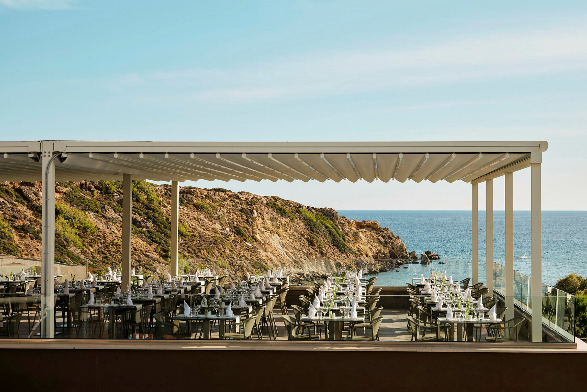 En udendørs restaurant med bord opdækket og udsigt over havet og klipper. (AI-genereret tekst)