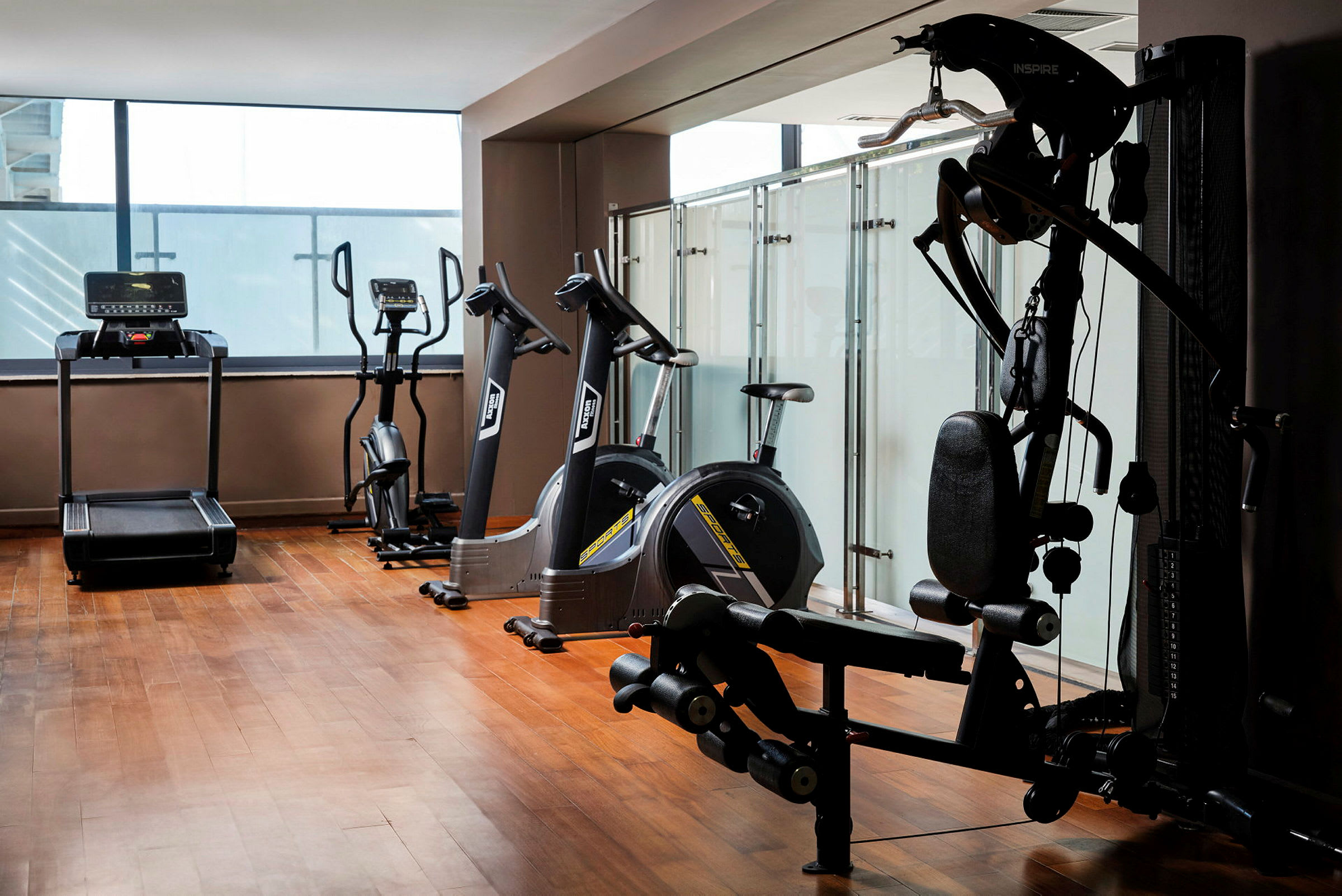 Et fitnesscenter med forskellige træningsmaskiner, herunder løbebånd, motionscykler og styrketræningsudstyr, beliggende i et rum med trægulv og glasvægge. (AI-genereret tekst)