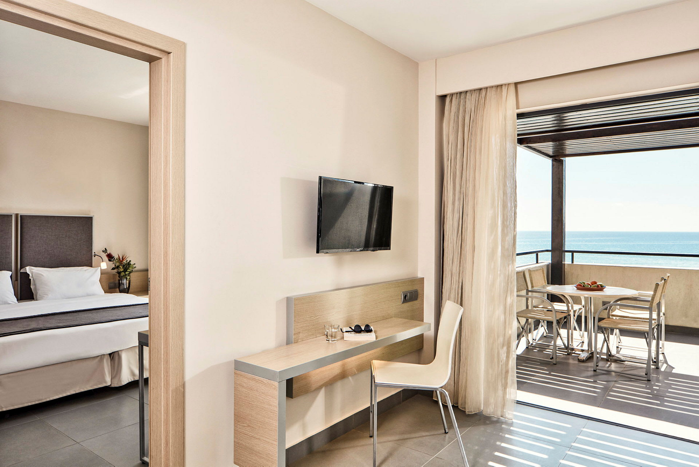Et hotelværelse med en seng, skrivebord, stol, vægmonteret TV og balkon med udsigt over havet. (AI-genereret tekst)