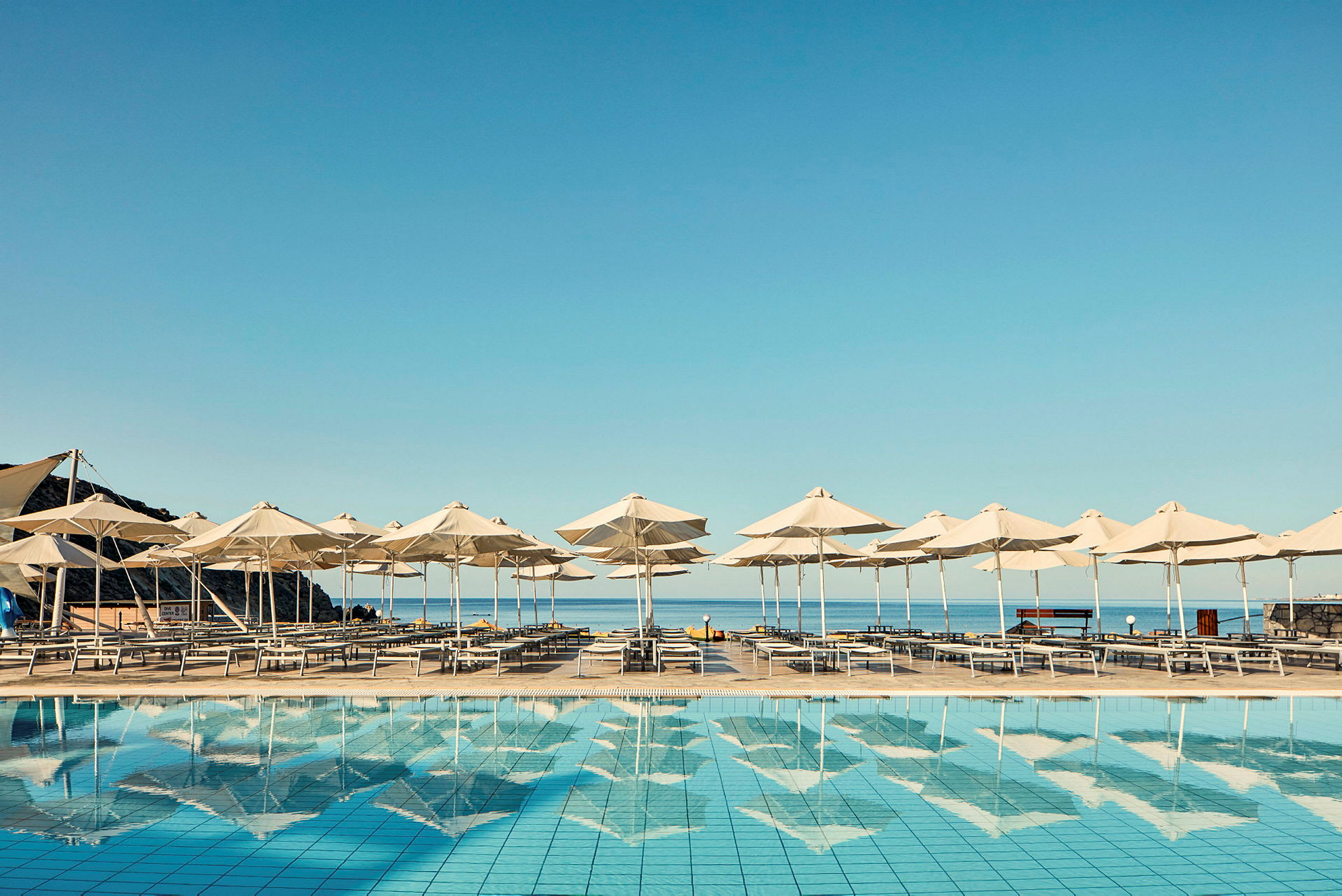 Liggestole og parasoller ved en swimmingpool med havet i baggrunden. (AI-genereret tekst)