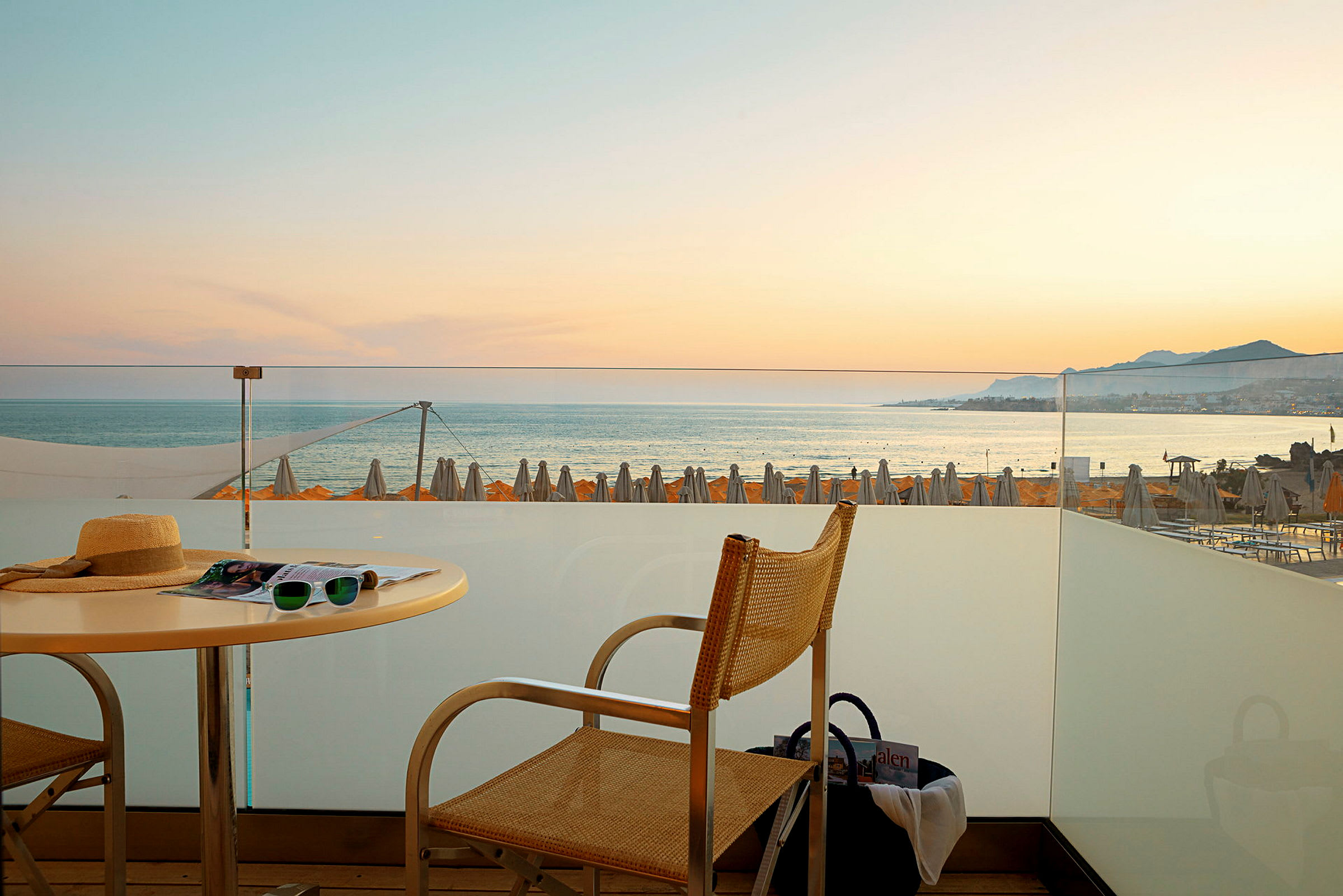 Udsigt fra en terrasse mod havet med solnedgang og strandparasoller i baggrunden. (AI-genereret tekst)