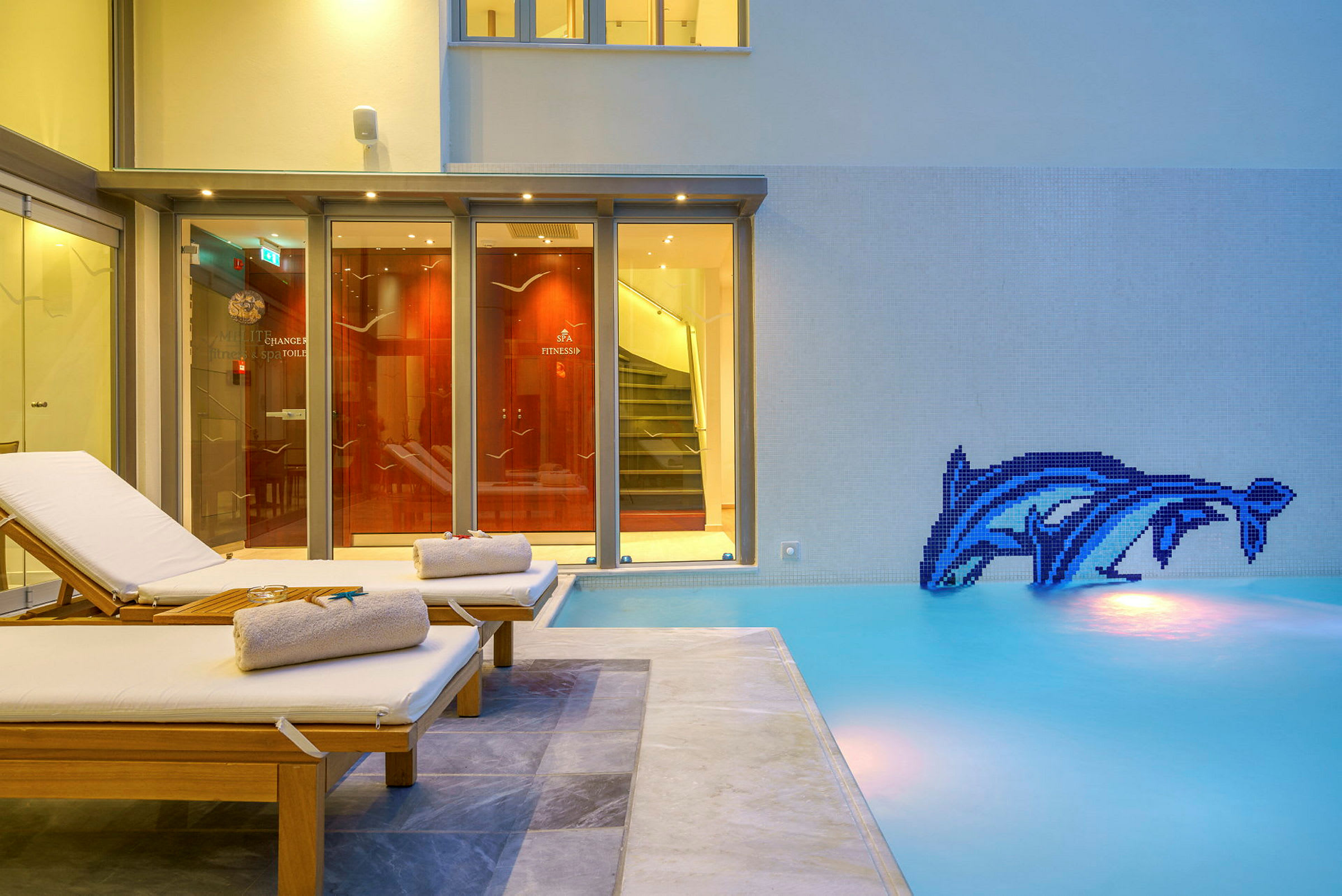 To liggestole med håndklæder ved en swimmingpool med en kunstnerisk mosaik af delfiner på væggen. (AI-genereret tekst)