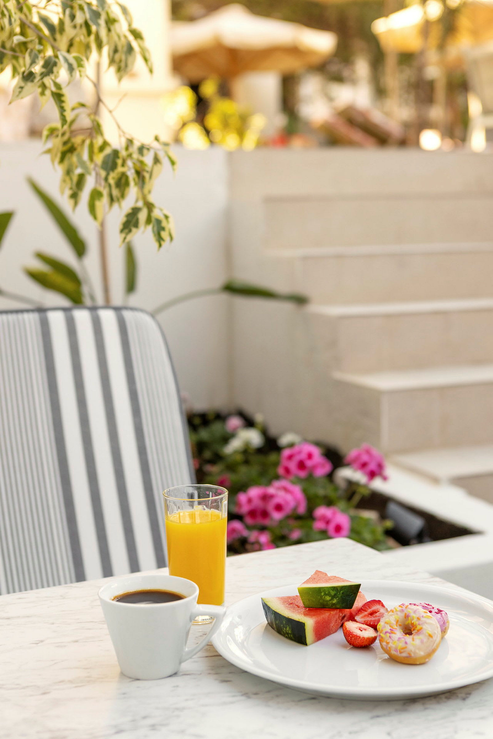 Et bord med en tallerken, der indeholder vandmelonstykker, jordbær og en donut ved siden af en kop kaffe og et glas appelsinjuice. I baggrunden ses blomster og trapper. (AI-genereret tekst)