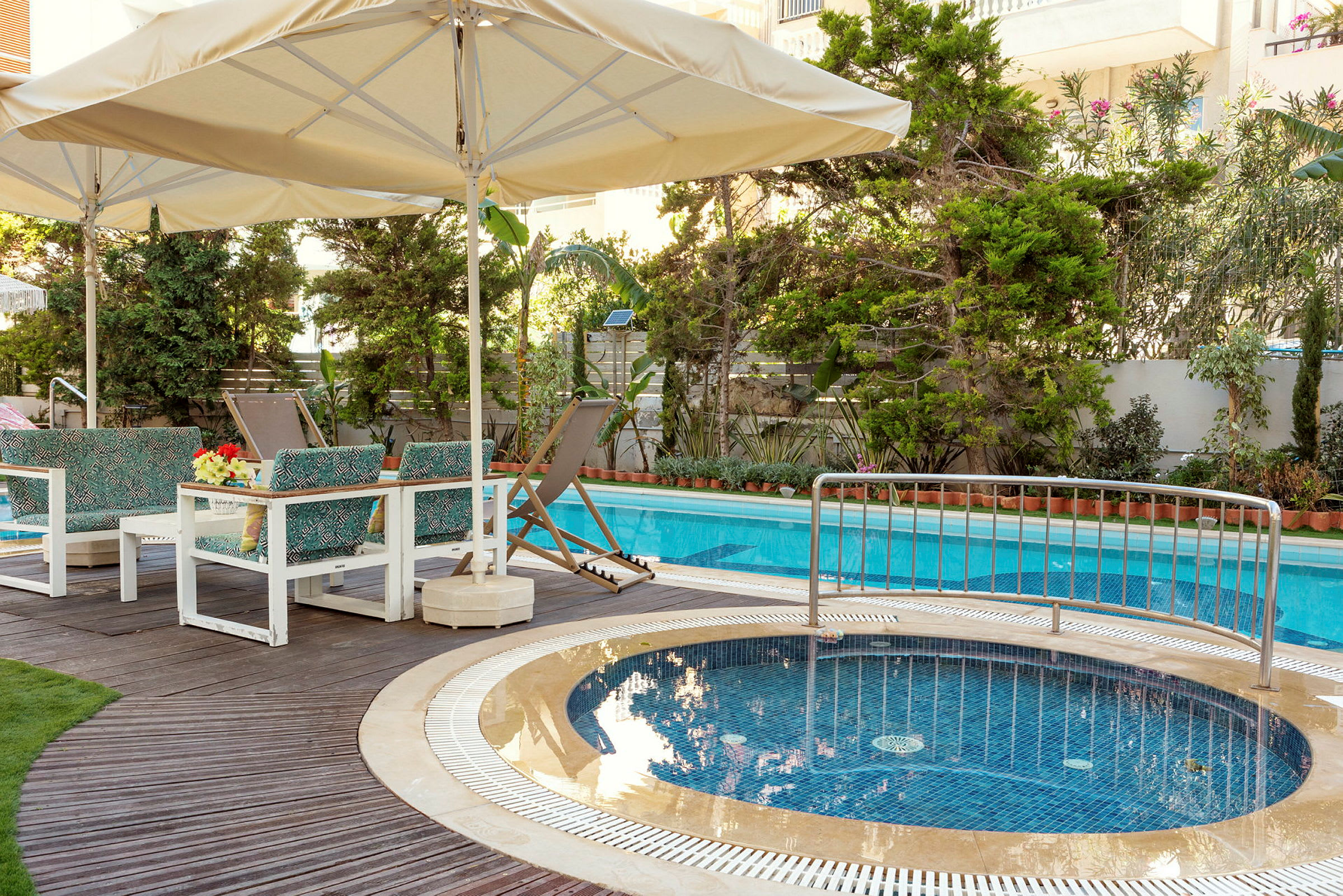Et poolområde med parasoller, liggestole og siddepladser ved en swimmingpool og en mindre jacuzzi. (AI-genereret tekst)