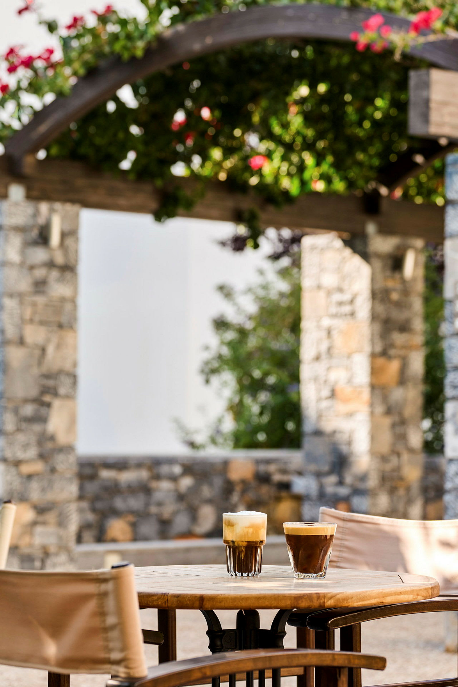 To glas med kaffe står på et bord udendørs, med en stenstruktur og blomstrende planter i baggrunden. (AI-genereret tekst)