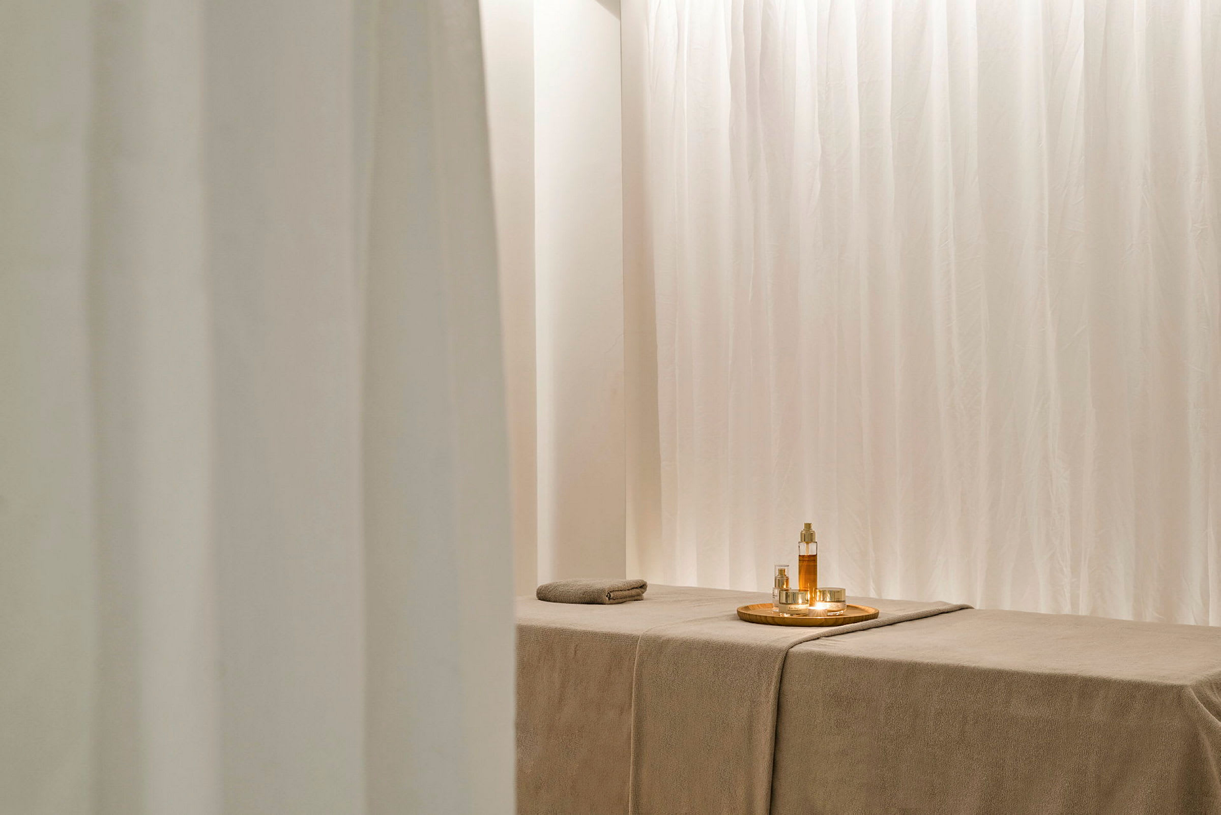 Et spa-rum med en massagebriks og lyse gardiner. På briksen er der en bakke med olier og små glas. (AI-genereret tekst)