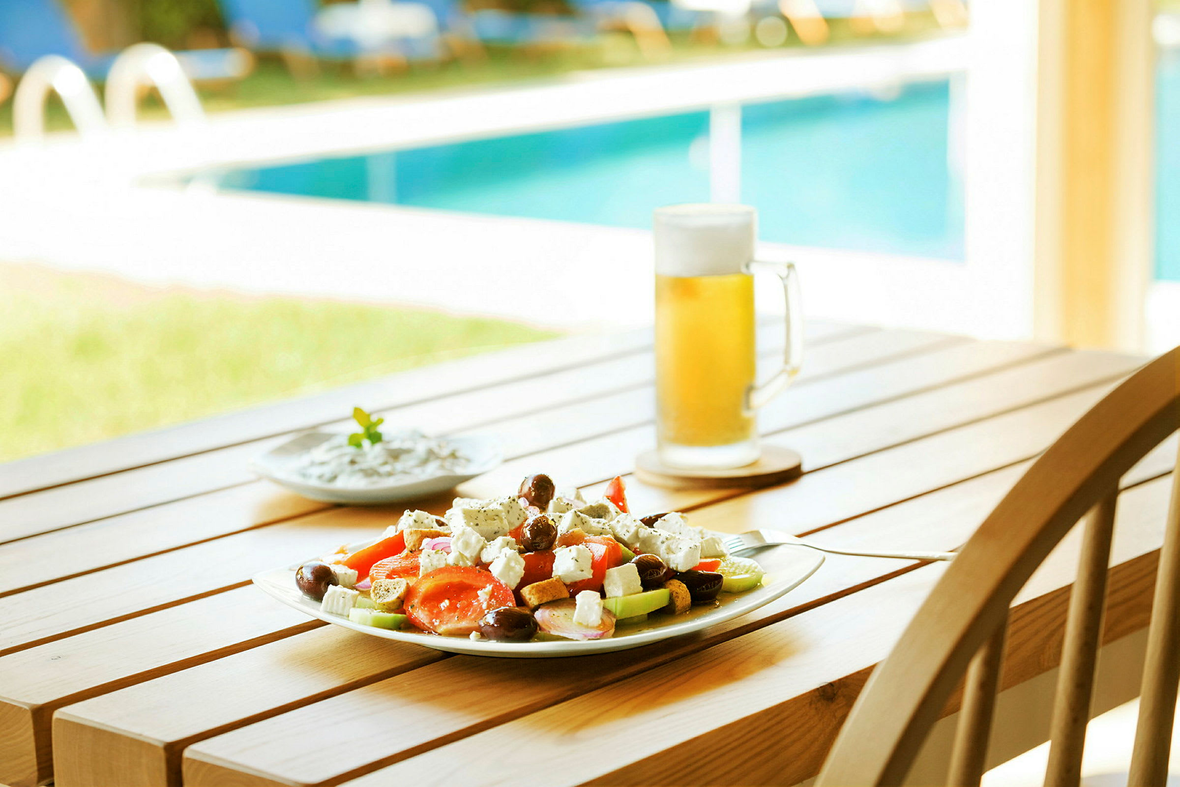 En tallerken med salat, et glas øl og en mindre skål står på et bord nær en pool. (AI-genereret tekst)