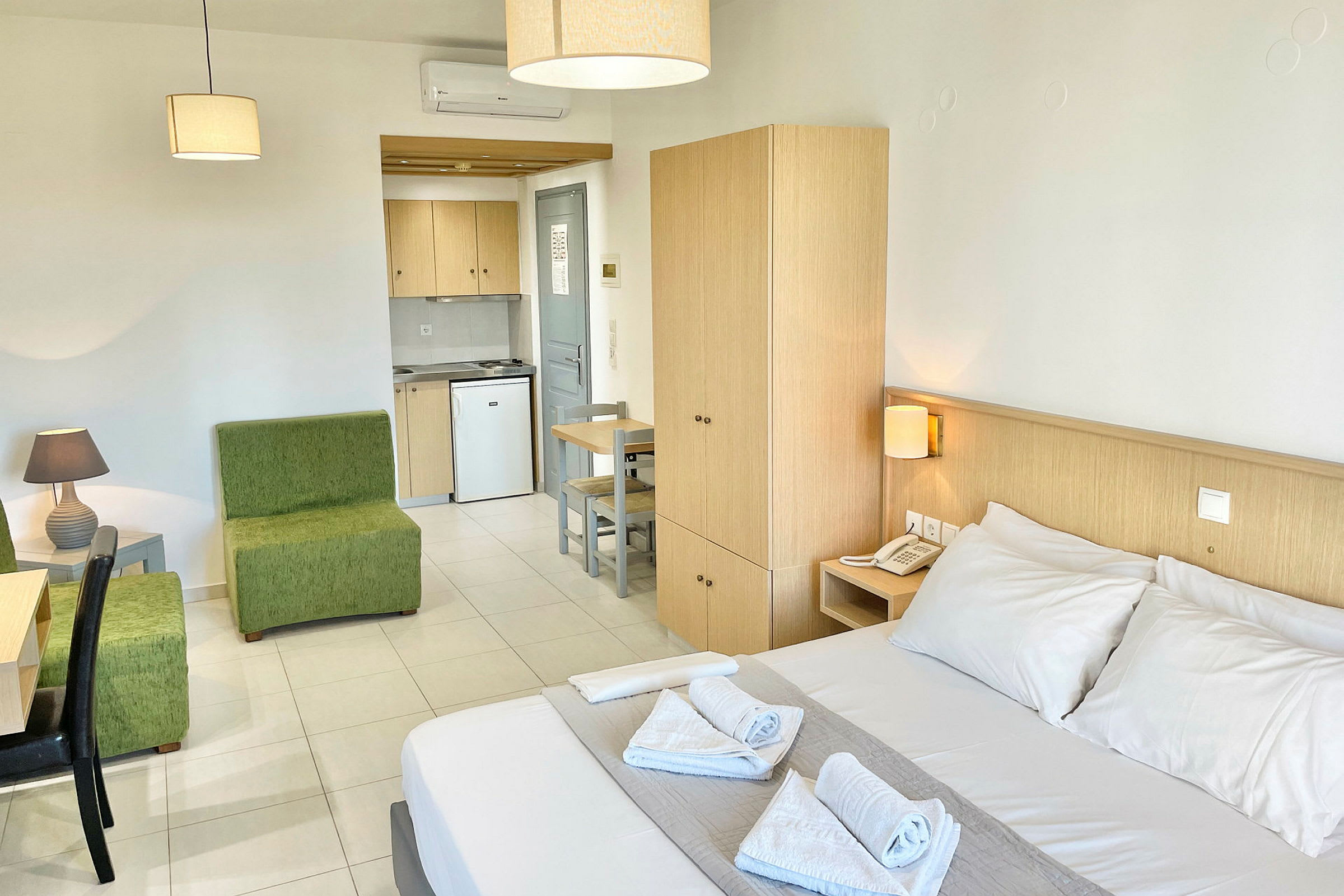 Et hotelværelse med dobbeltseng, grøn siddegruppe, skrivebord, garderobe og et tekøkken. (AI-genereret tekst)