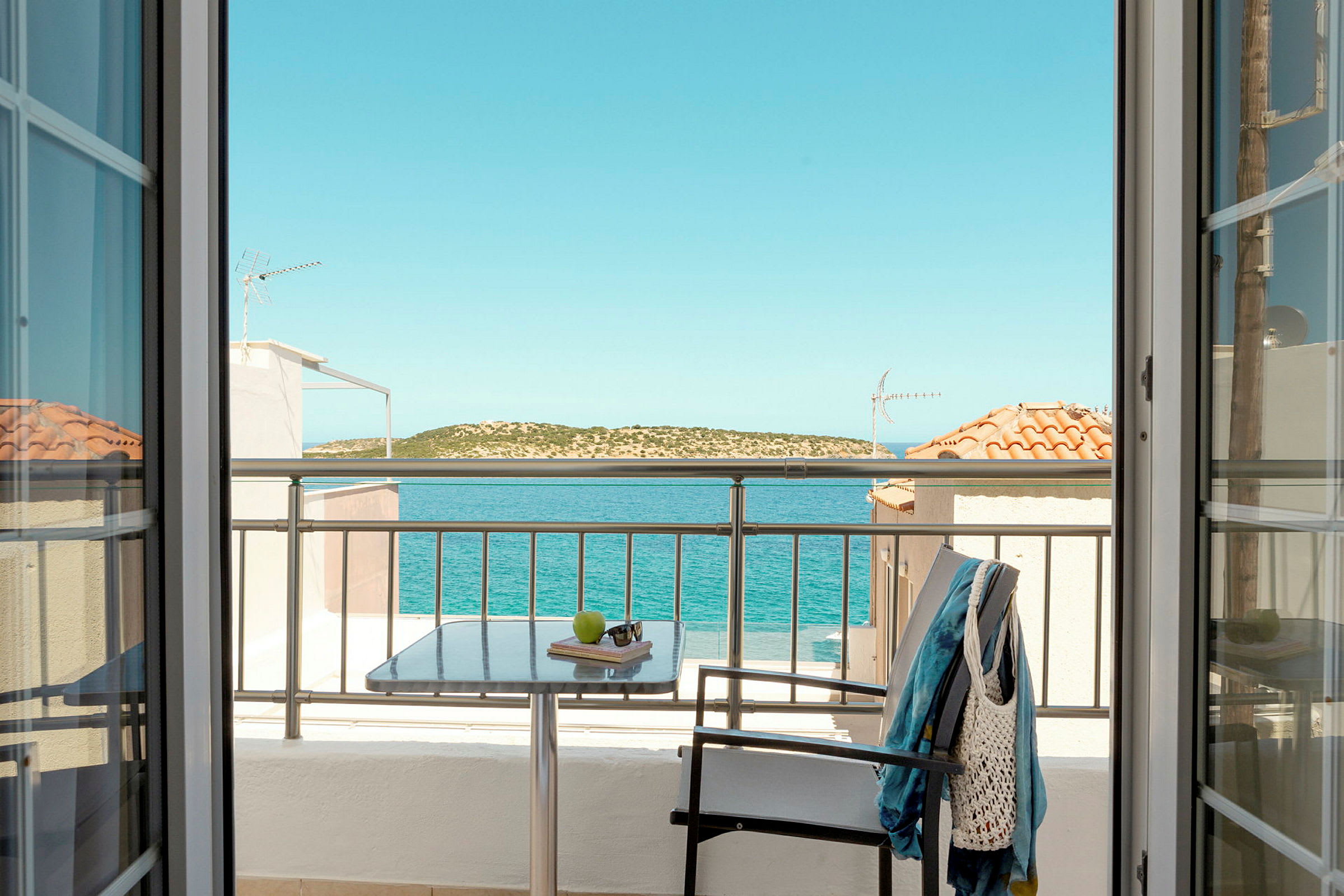 Udsigt fra et værelse gennem døre mod en balkon med bord, stol og tøj, samt hav og landskab i baggrunden. (AI-genereret tekst)