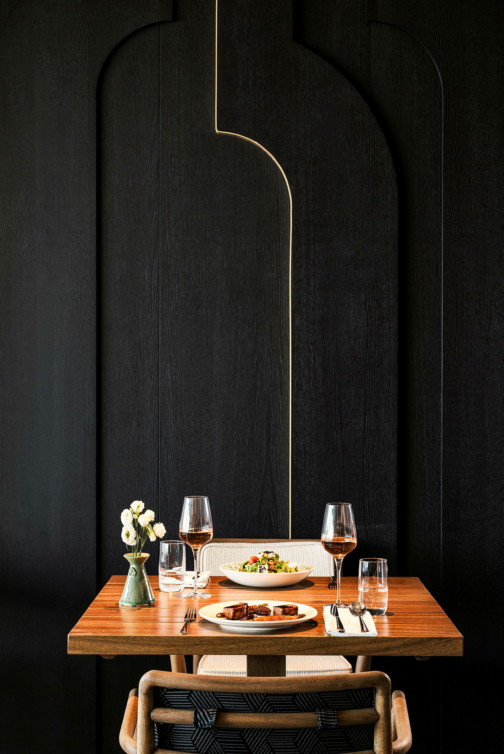 Et dækket bord med en træoverflade, til to personer, to glas vin, to tallerkener med mad og en vase med blomster. (AI-genereret tekst)