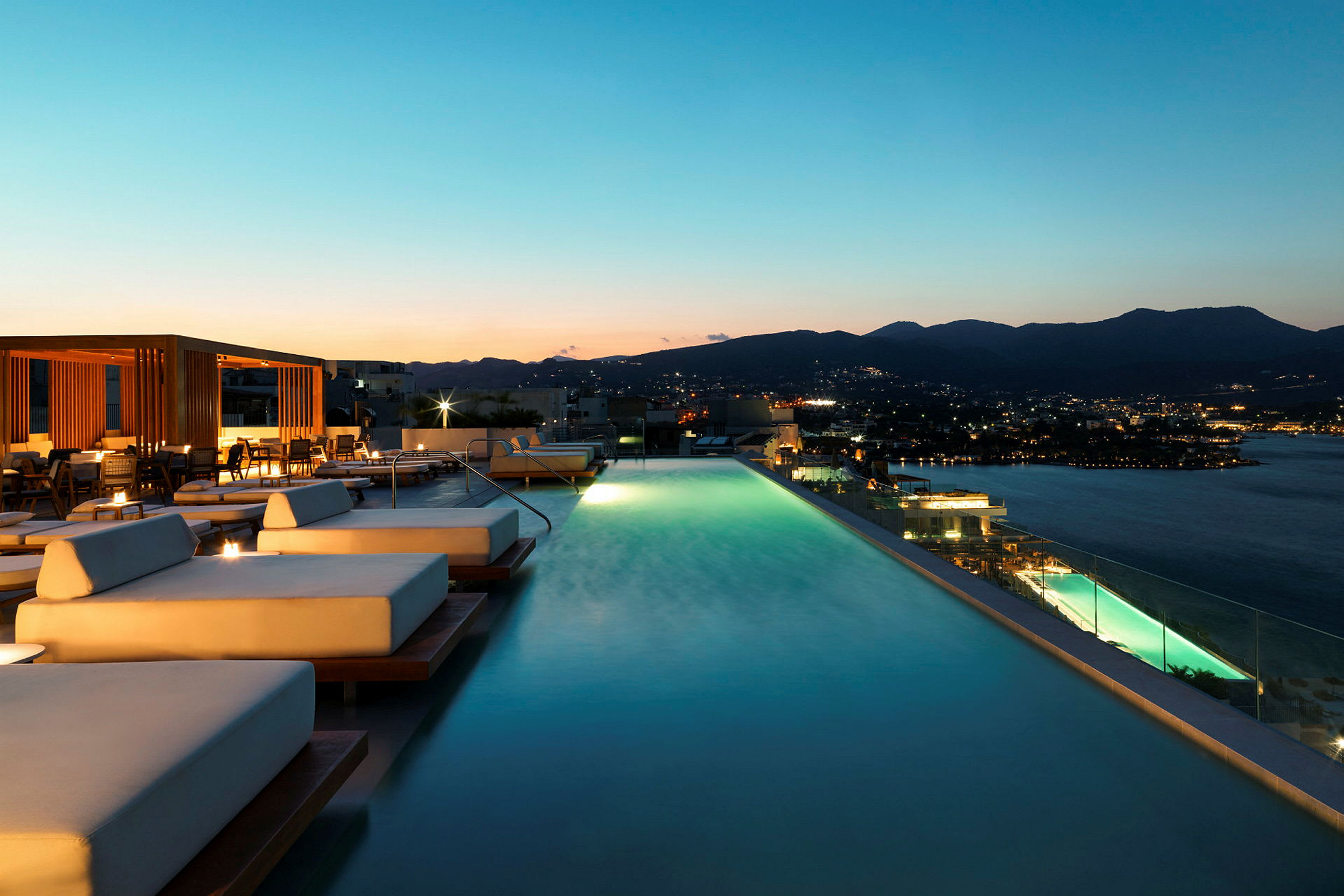 Infinitypool med solsenge ved solnedgang, med udsigt over byen og havet. (AI-genereret tekst)