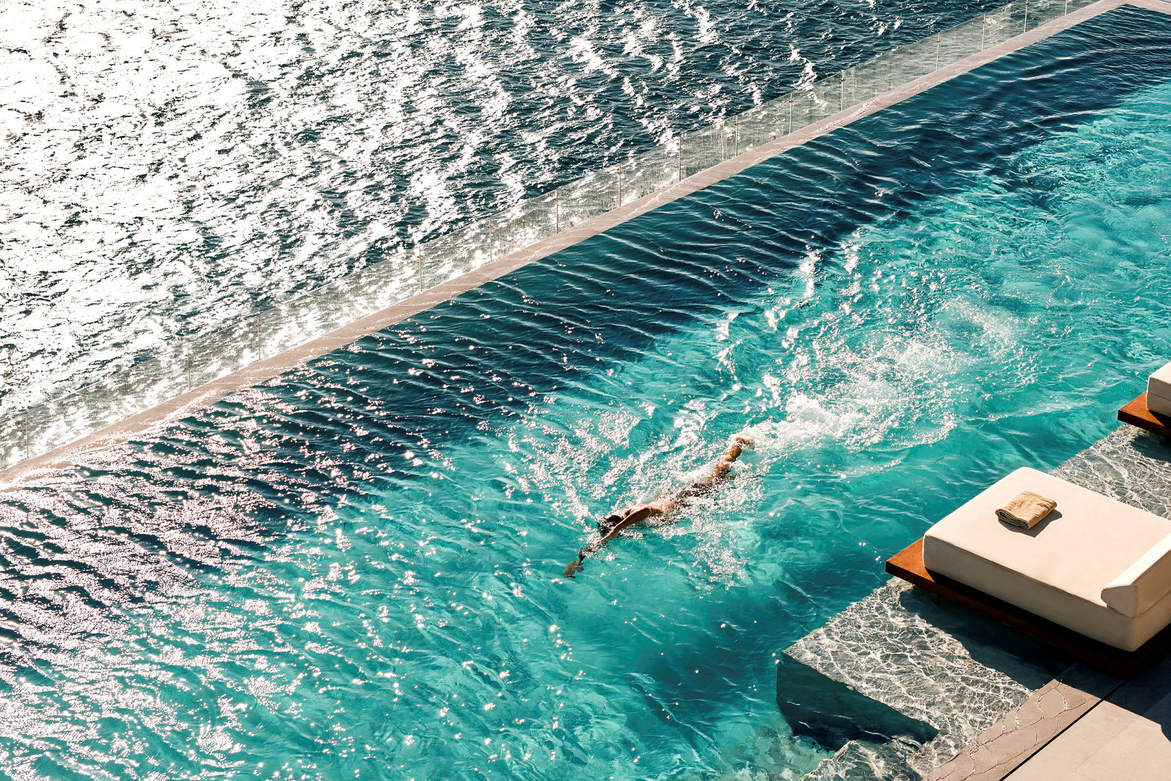 En person svømmer i en pool ved havet, med liggestole i forgrunden. (AI-genereret tekst)