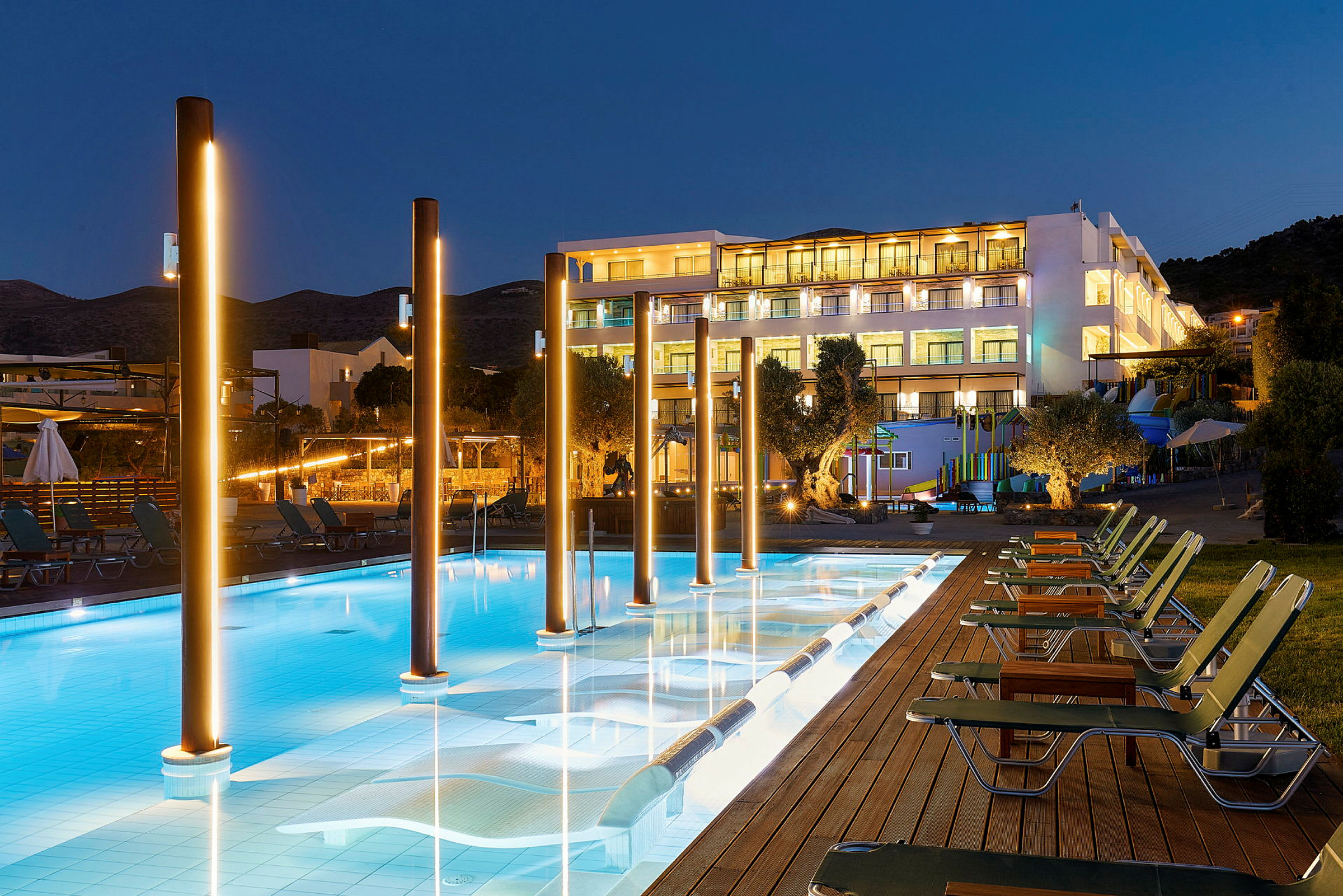 En oplyst hotelfacade bag en pool med stole ved siden, fotograferet om aftenen. (AI-genereret tekst)