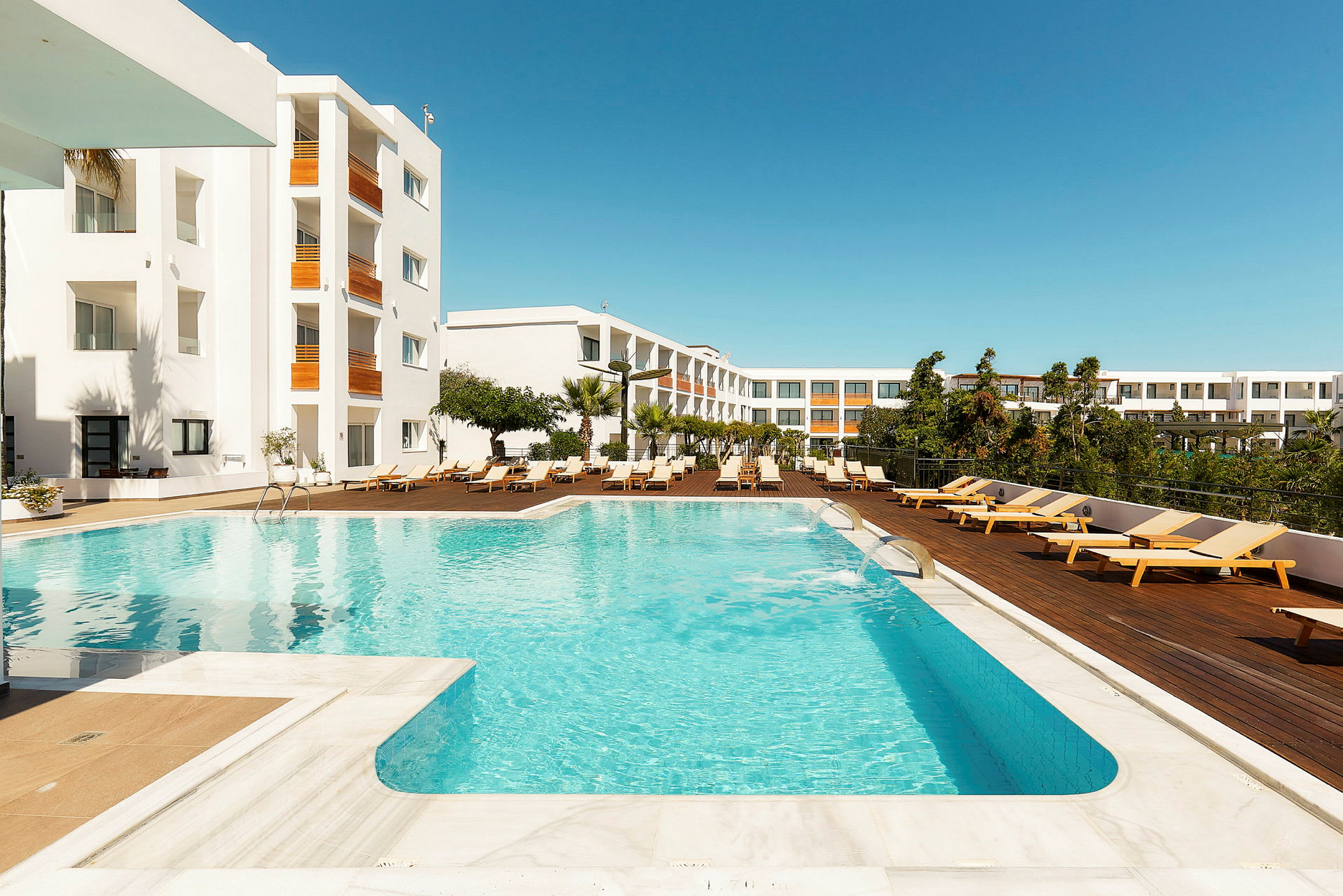 Swimmingpool foran et hotel med hvide facader og liggestole på en træterrasse. (AI-genereret tekst)