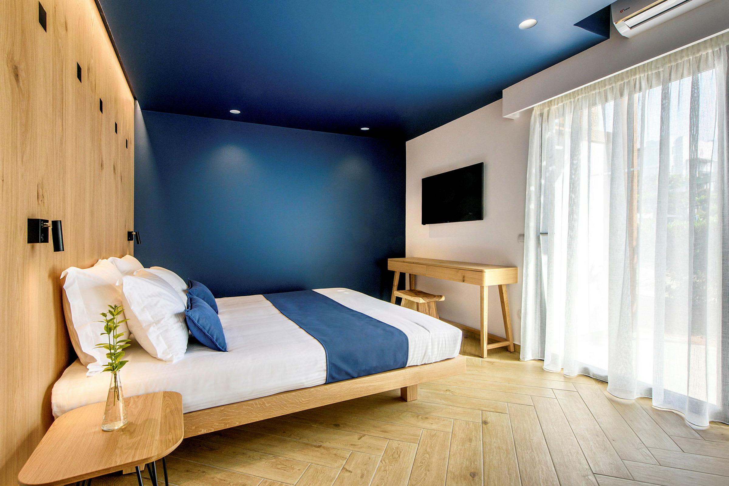 Et moderne hotelværelse med blå vægge, en seng med blå sengetæppe og træmøbler. (AI-genereret tekst)