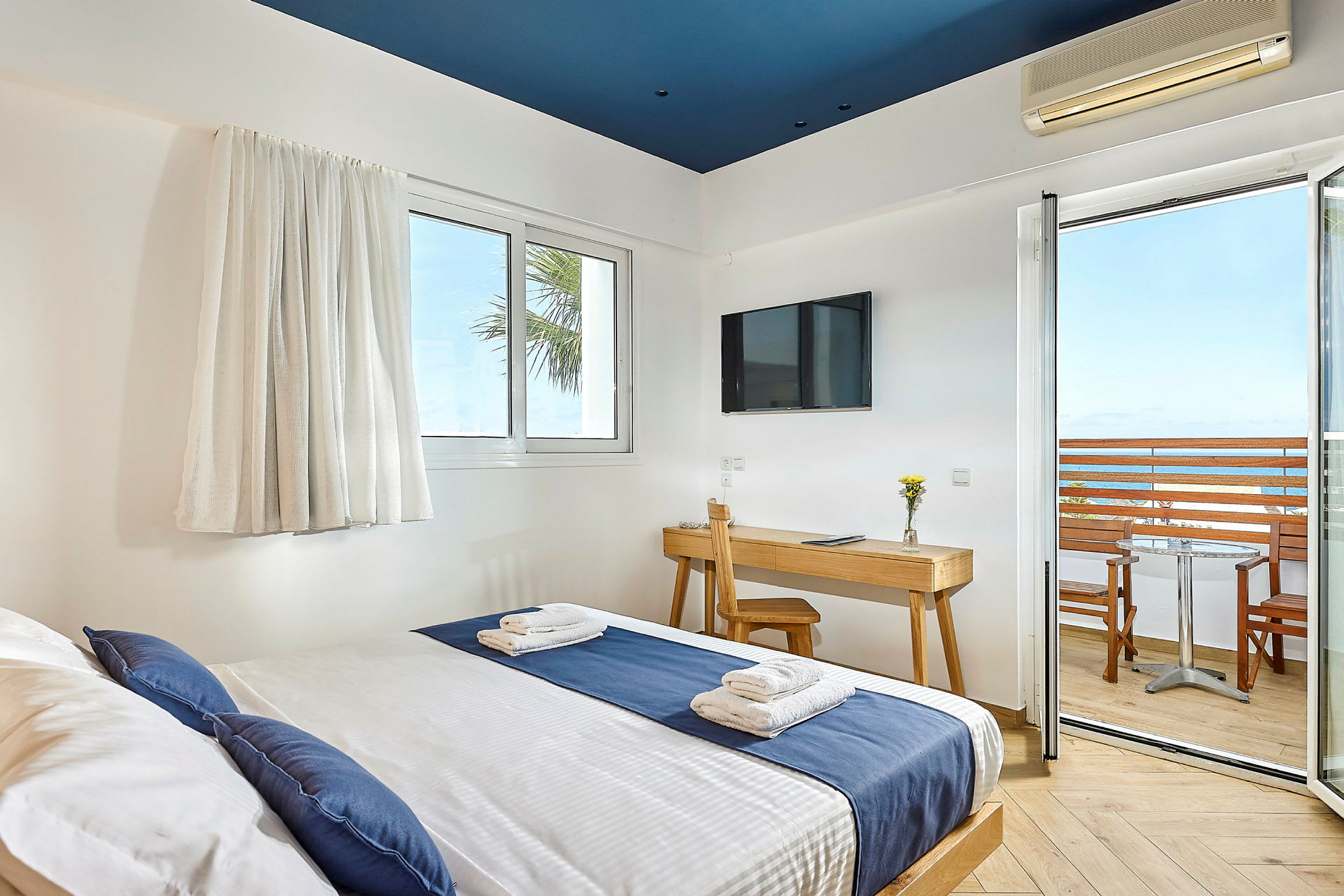 Hotelværelse med seng beklædt med blåt og hvidt sengetøj, skrivebord med stol, TV på væggen og balkon med udsigt over havet. (AI-genereret tekst)