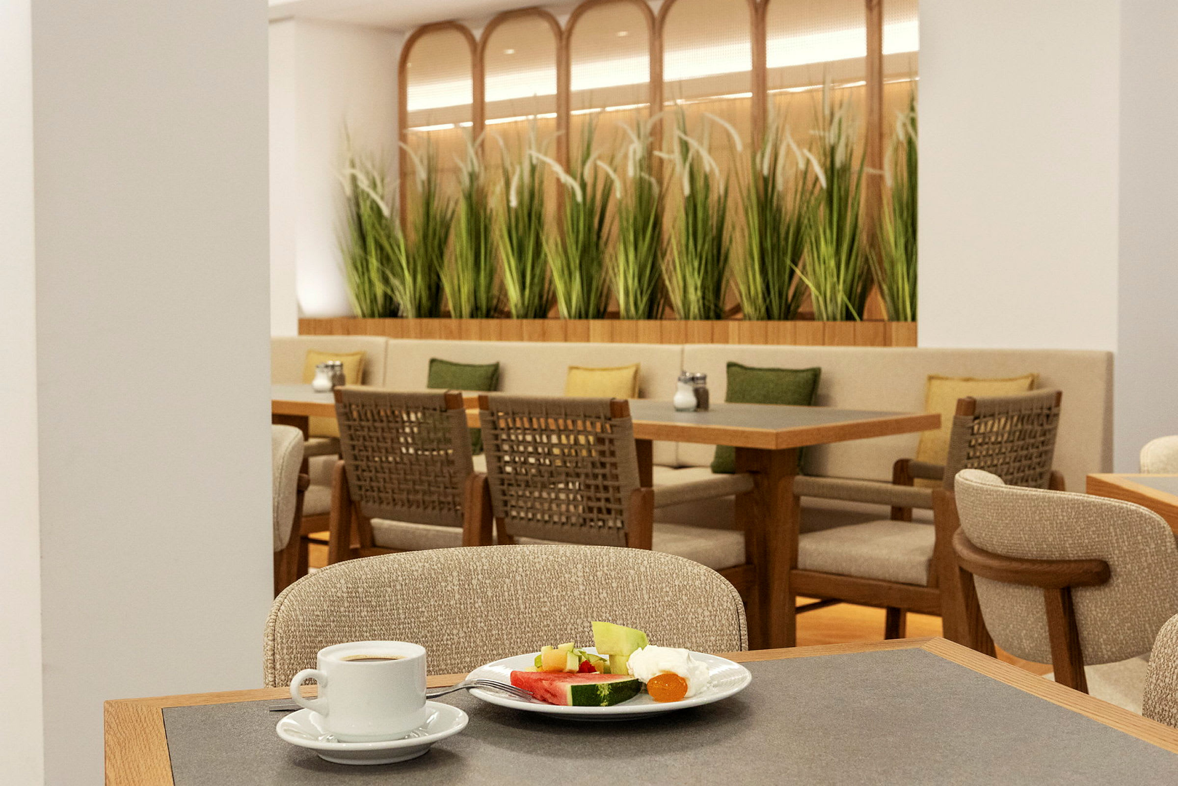 Billede af en restaurant med borde, stole og grønne planter som dekoration. I forgrunden ses en kaffekop og en tallerken med frugt. (AI-genereret tekst)