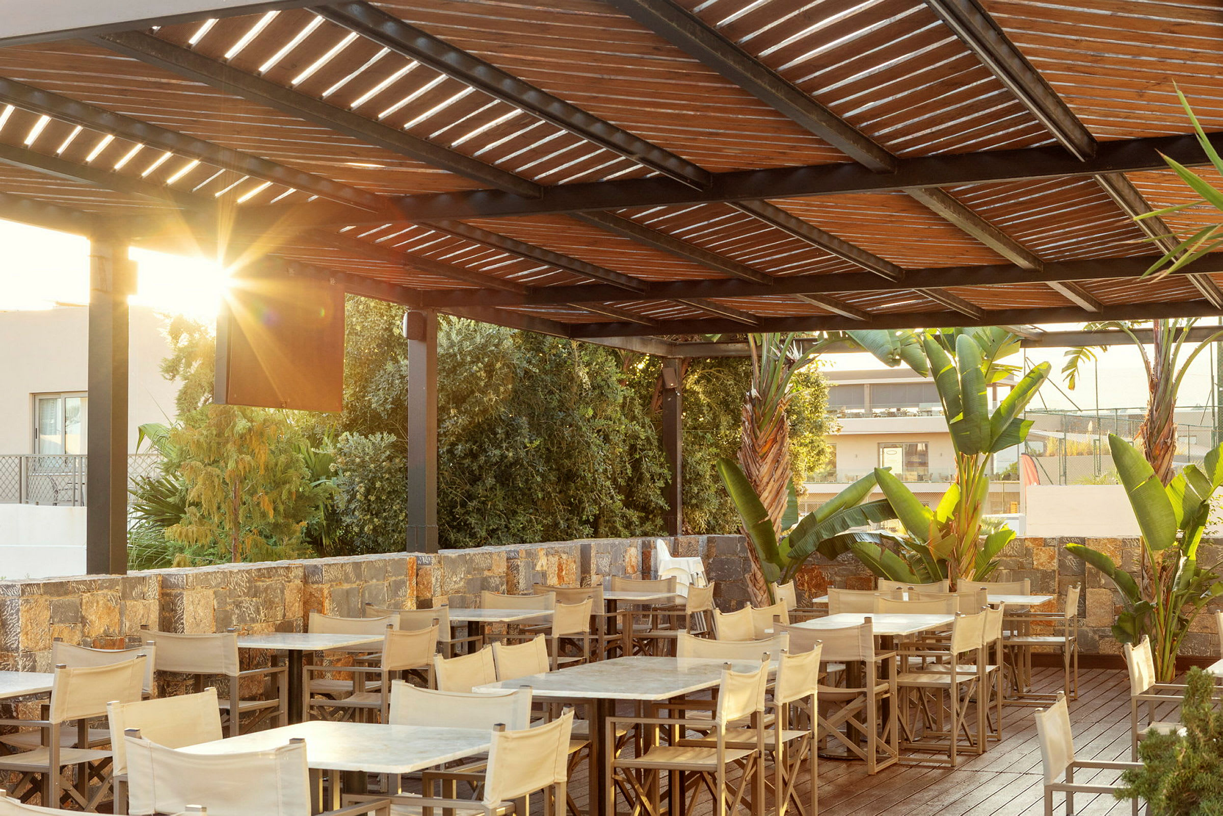 En udendørs restaurant med borde, stole, en træpergola og omkringliggende vegetation. (AI-genereret tekst)