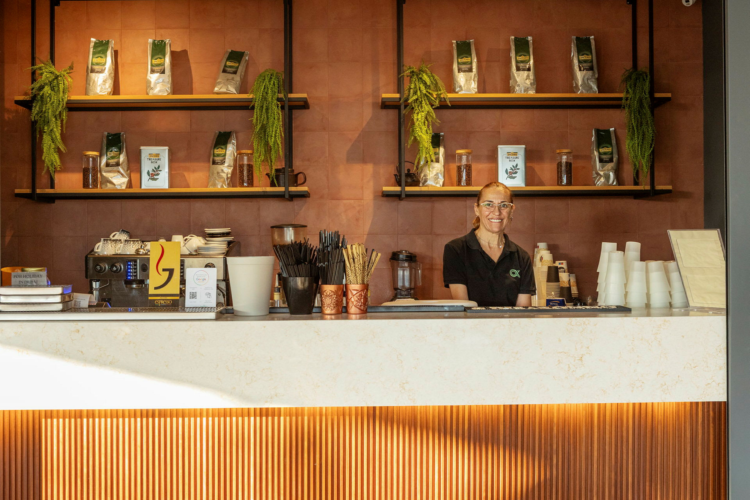 En person står bag en disk i en café, omgivet af hylder med kaffeprodukter og dekorationer. (AI-genereret tekst)