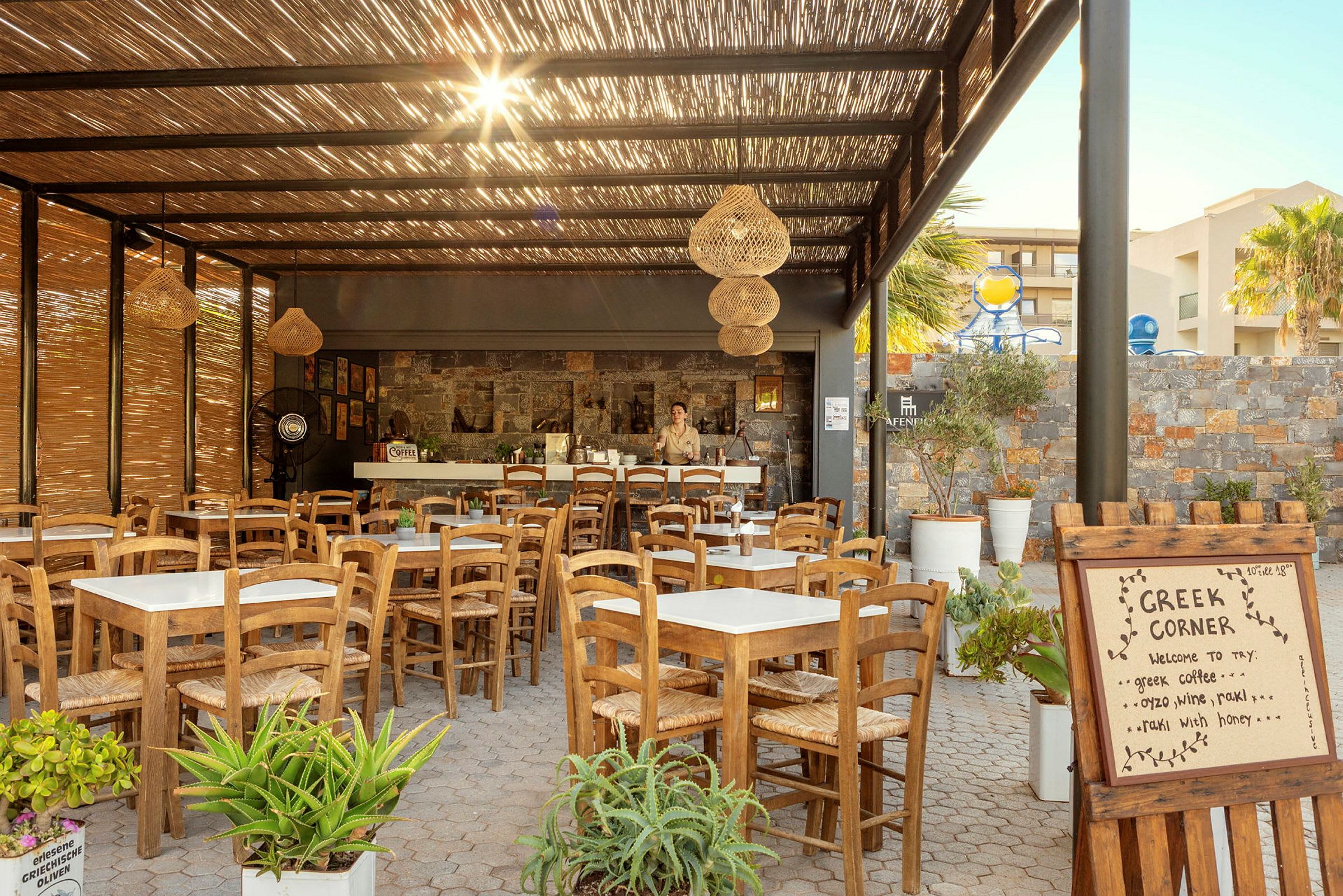 Dette billede viser en udendørs restaurant med træborde og stole under et tag af bambus samt et skilt med teksten "Greek Corner". (AI-genereret tekst)