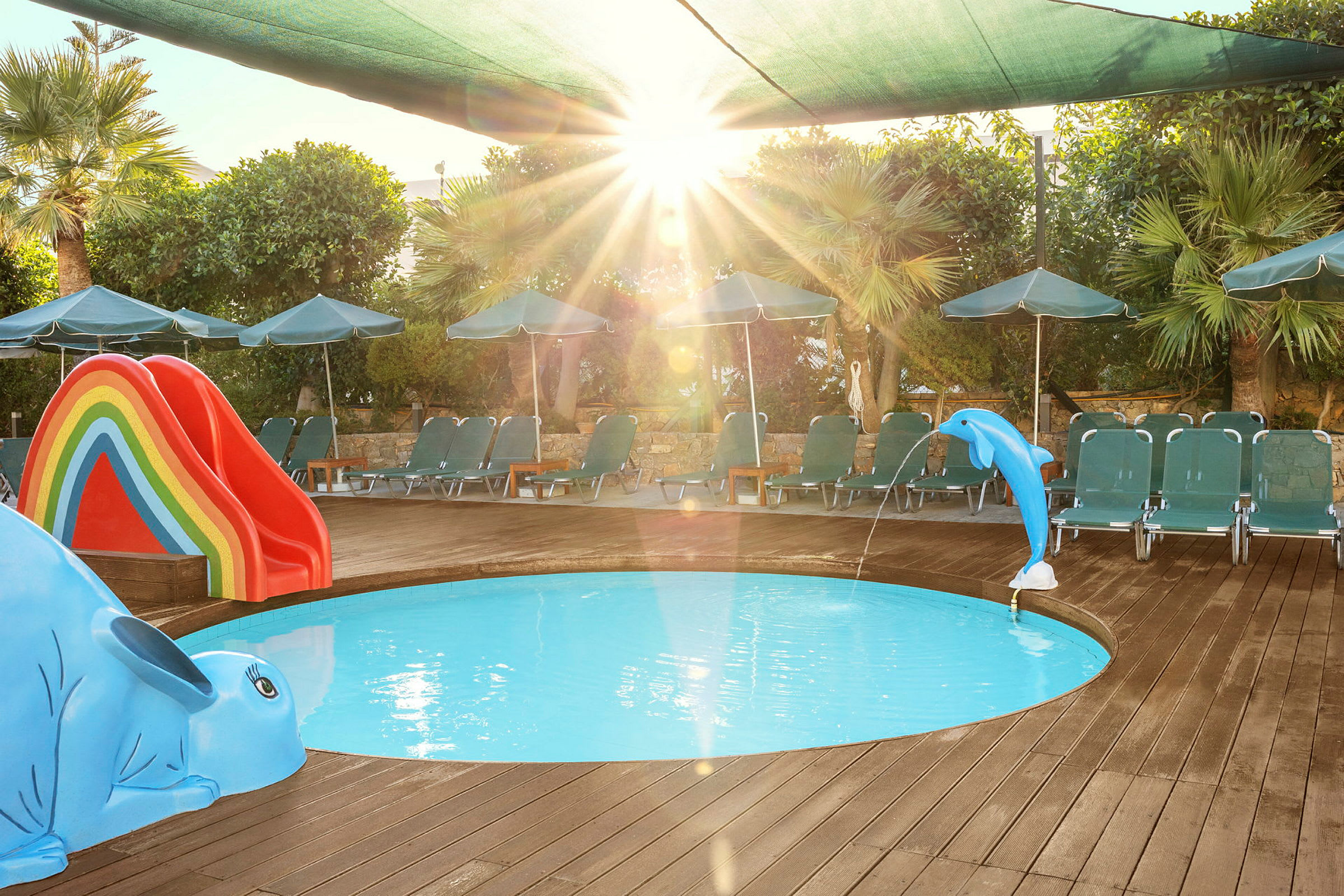 En lille, rund børnepool omgivet af en træterrasse. I poolen er der en blå delfinfigur, der sprøjter vand, og en rutsjebane i regnbuens farver. Liggestole og parasoller står omkring poolen. (AI-genereret tekst)