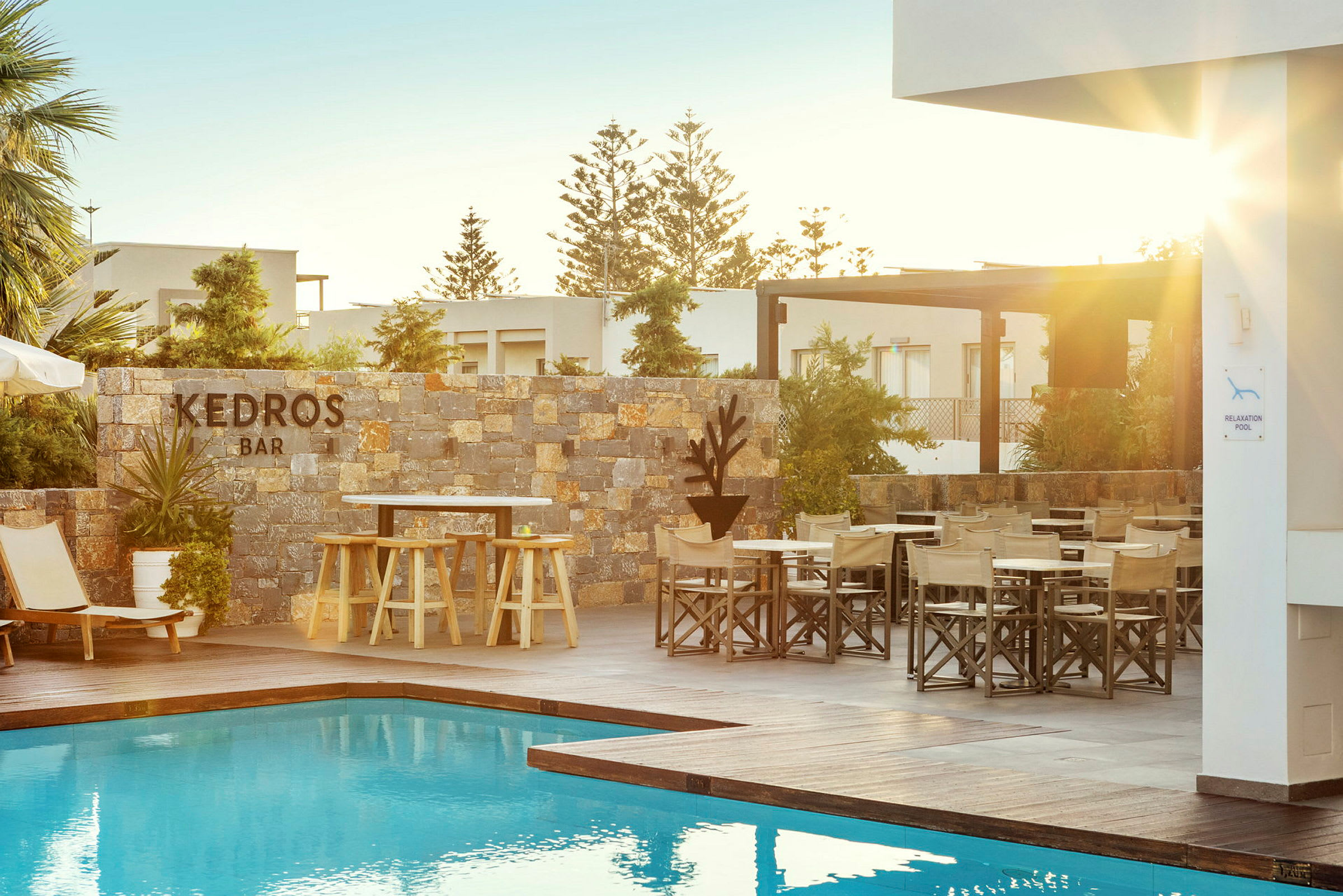 En udendørs pool med solsenge og borde ved siden af en stenmur mærket "Kedros Bar". Solen skinner over området. (AI-genereret tekst)