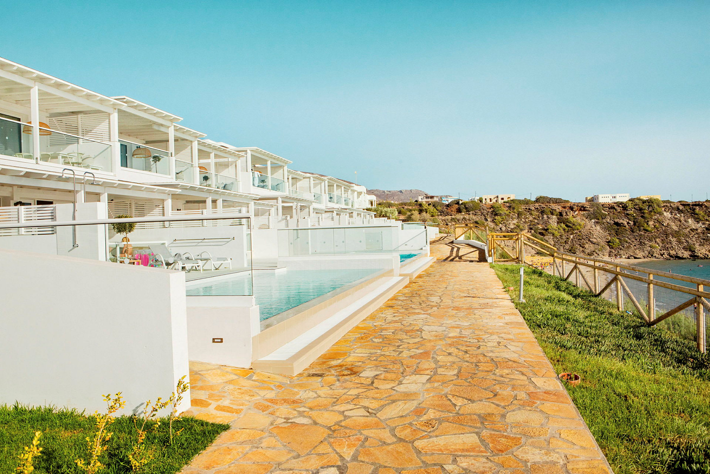 Billede af et moderne feriested med hvid arkitektur, en gangsti, swimmingpools og udsigt til kysten. (AI-genereret tekst)