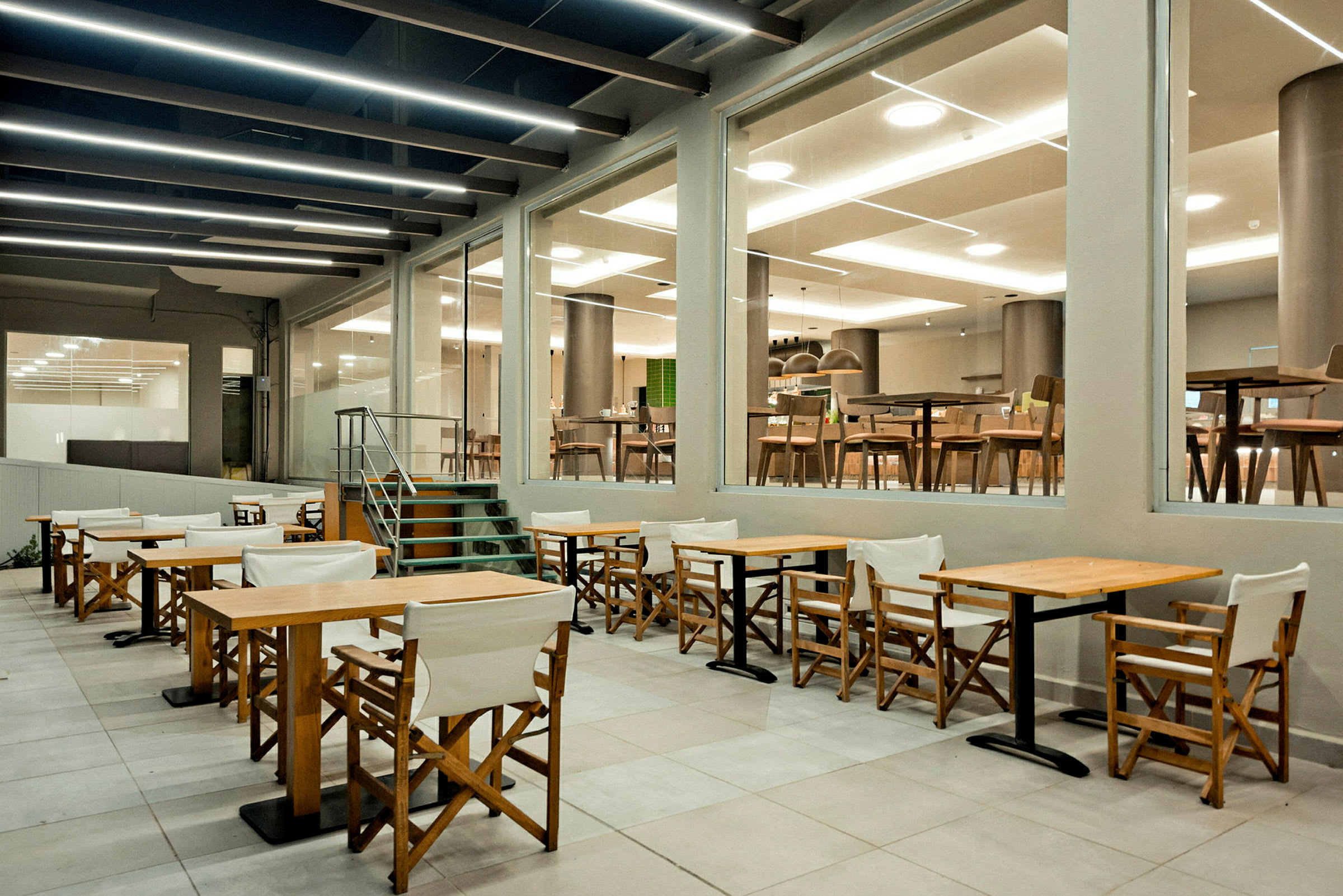 Udendørsområde med træborde og stole foran en glasfacade, der viser en lys restaurantinteriør. (AI-genereret tekst)