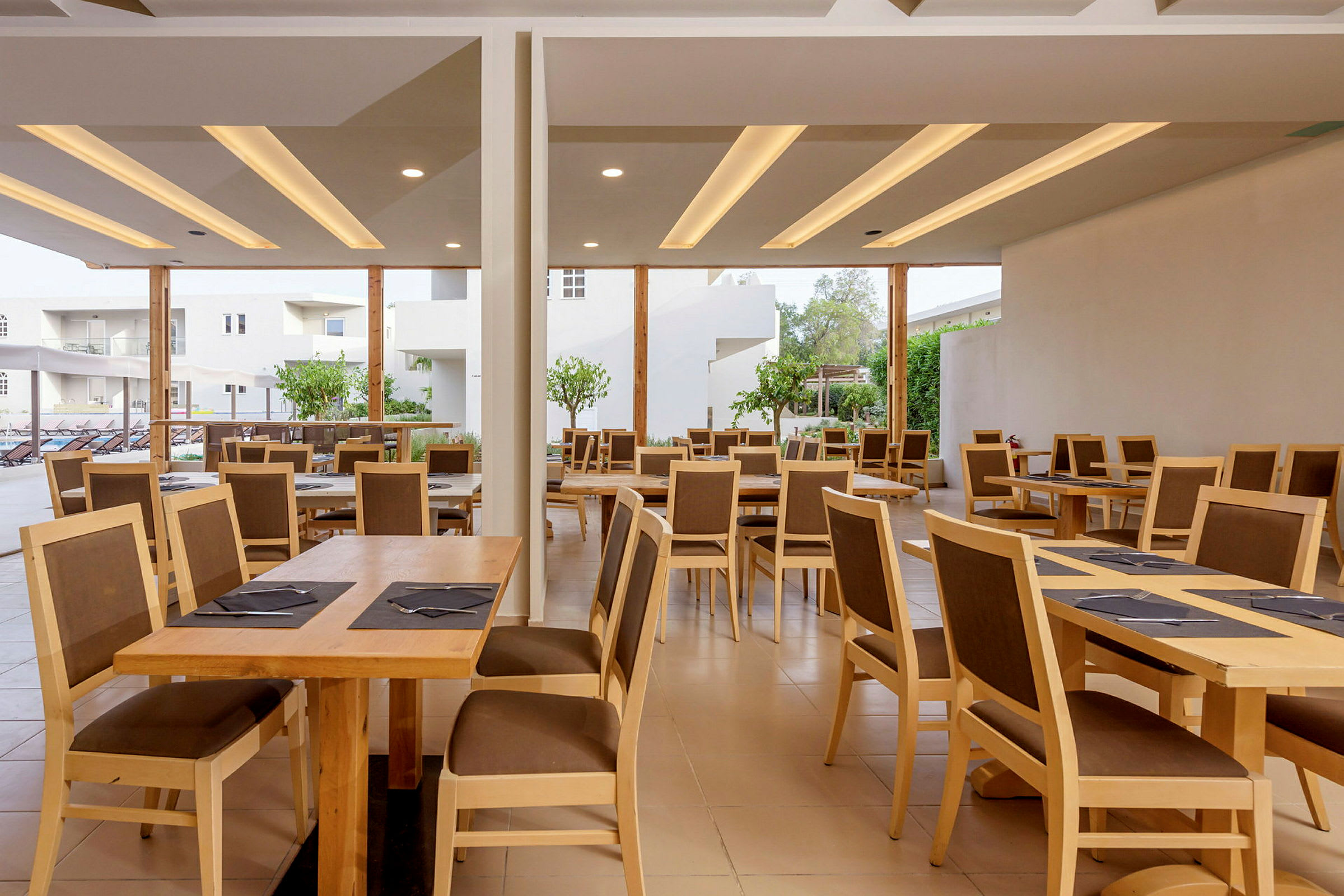 Restaurant med træborde og stole, åben mod en udendørs terrasse. (AI-genereret tekst)