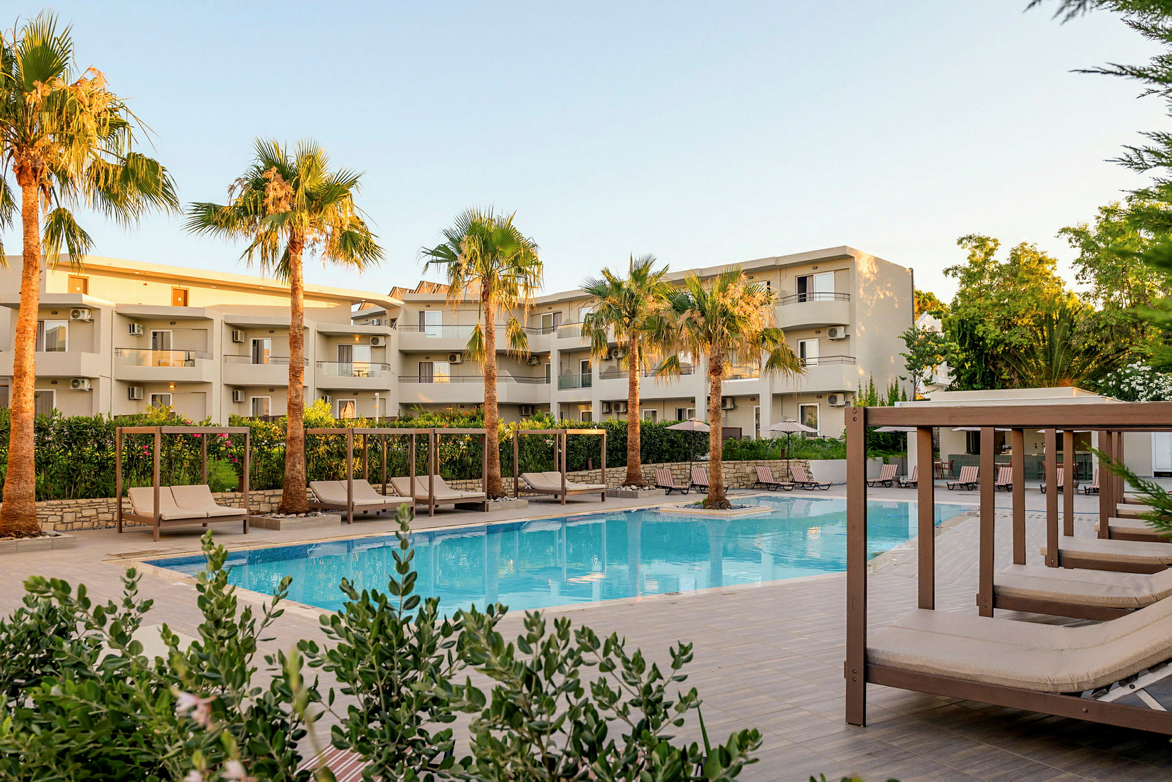 Billede af en moderne hotelbygning med altaner omgivet af grønne palmer. Foran bygningen er der en pool og flere solsenge under skyggetage. (AI-genereret tekst)