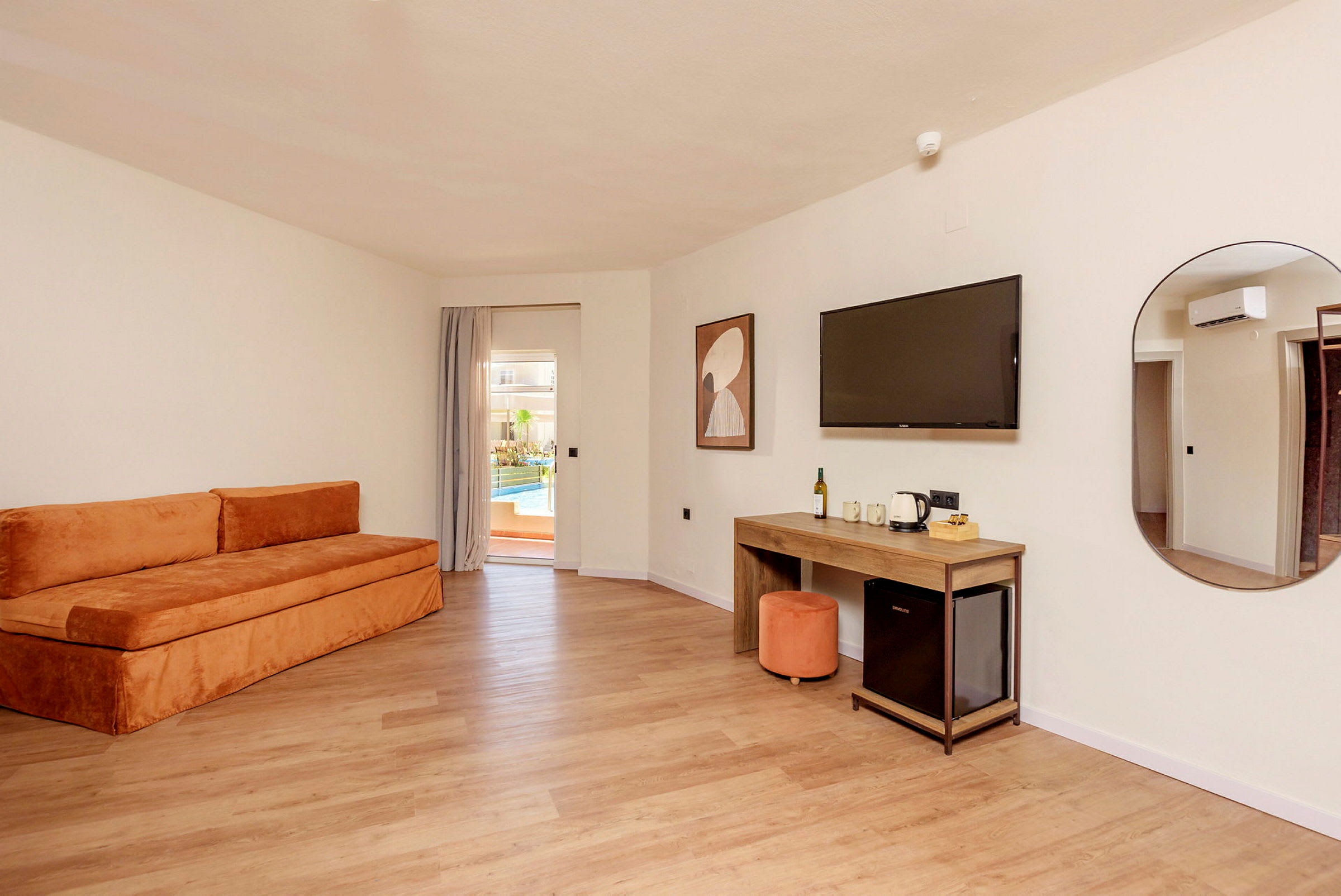 Et hotelværelse med en orange sofa, et skrivebord, et vægmonteret TV og en dør til en terrasse med pool. (AI-genereret tekst)