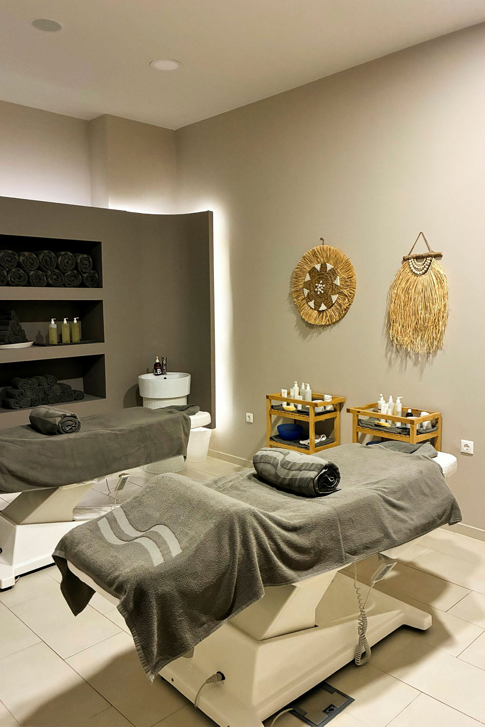 Et spa-rum med to behandlingsborde dækket med grå frottéhåndklæder. Væggene er dekoreret med naturinspirerede kunstværker, og der er hylder med mørke håndklæder og produkter i baggrunden. (AI-genereret tekst)