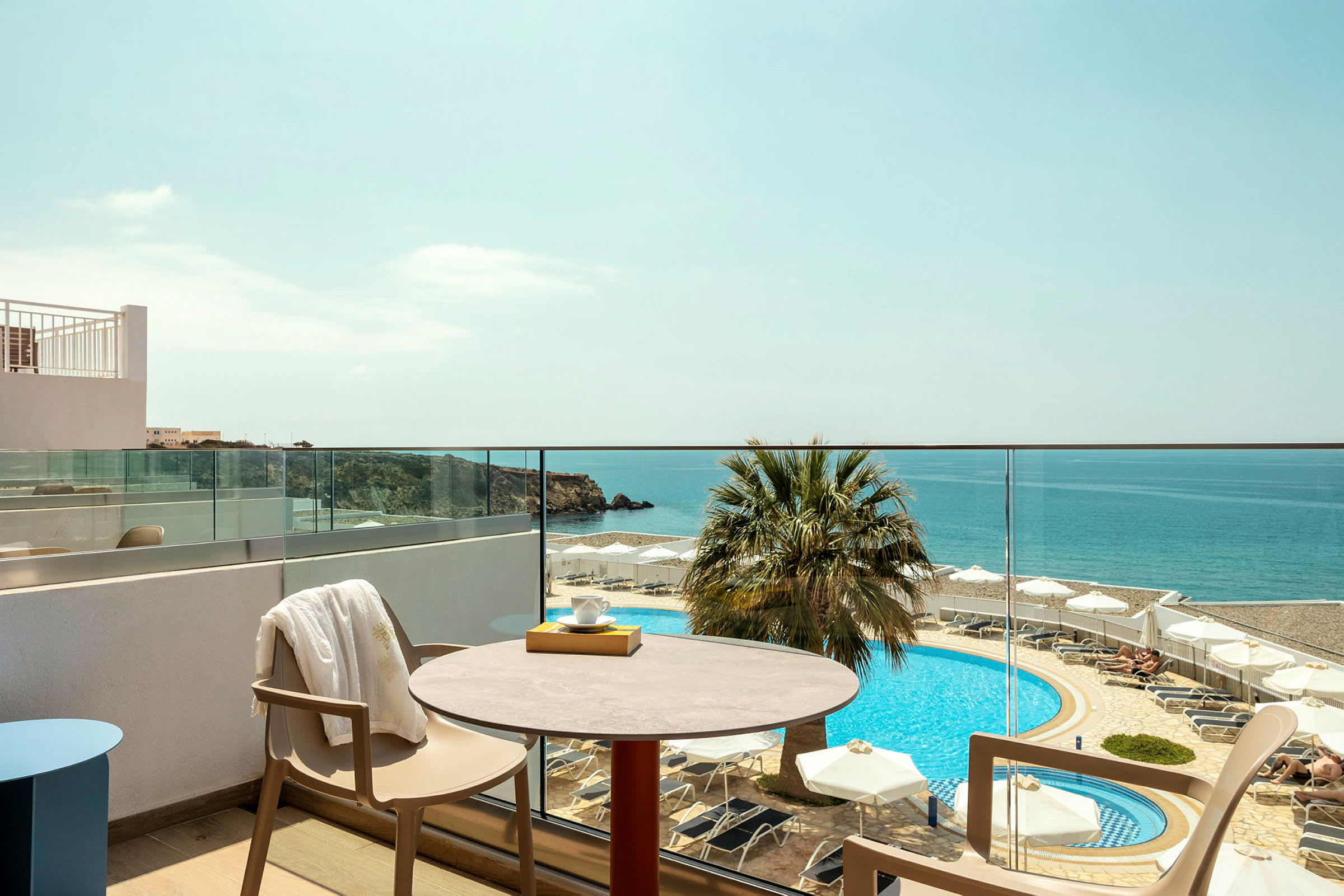 Udsigten fra en balkon mod havet, hvor en pool omgivet af liggestole og parasoller kan ses. (AI-genereret tekst)