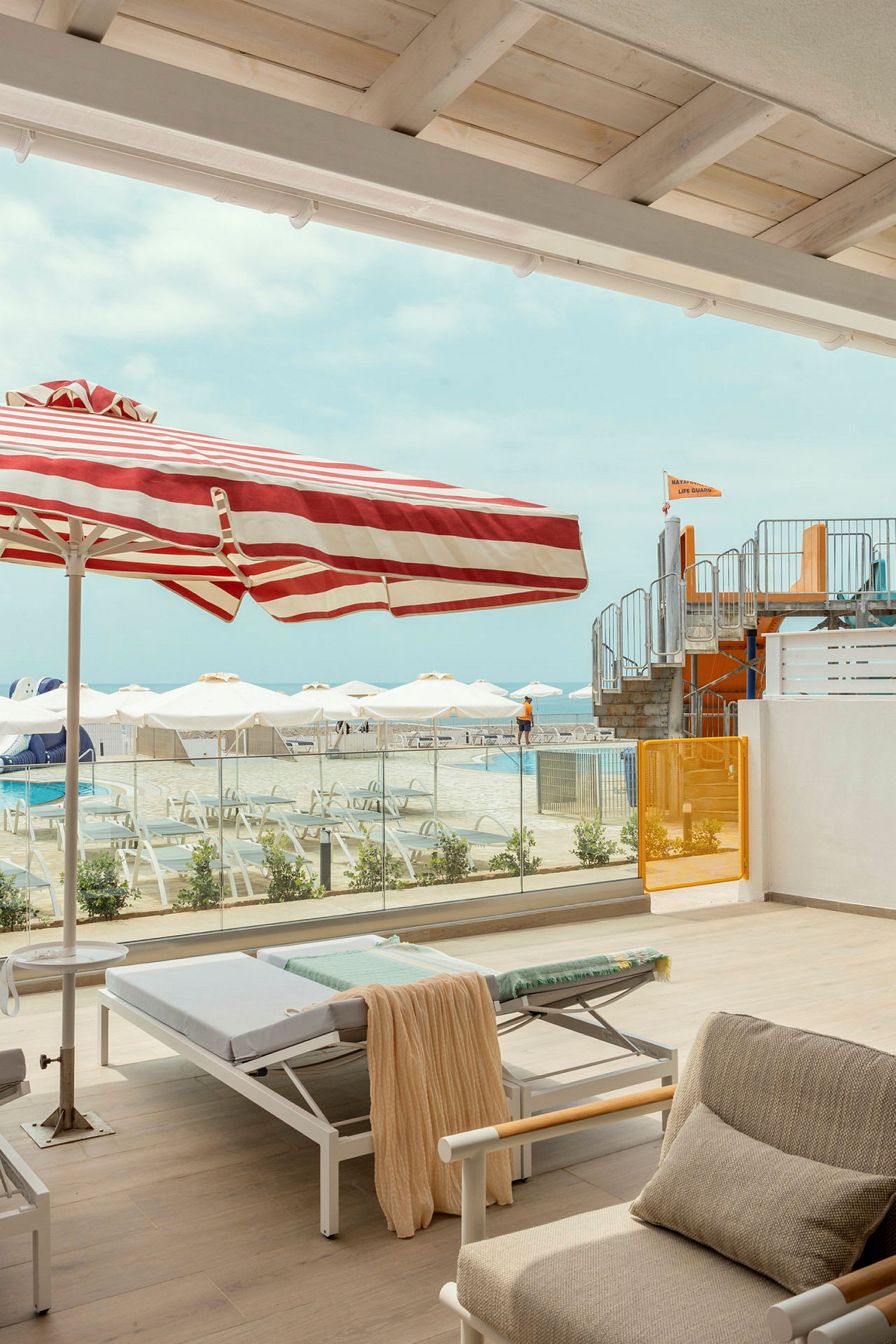 Udsigt fra en terrasse med liggestole og en stribet parasol mod et poolområde med parasoller og vandrutsjebaner i baggrunden. (AI-genereret tekst)