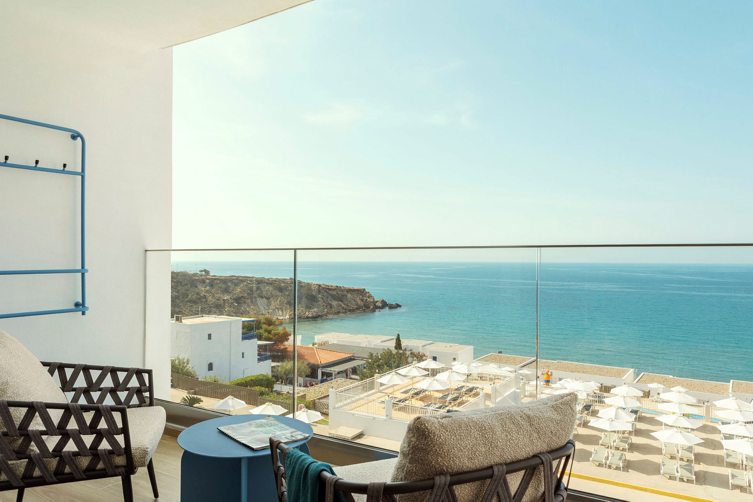 Udsigt fra en balkon med moderne møbler mod havet, klipper og bygninger ved kysten. (AI-genereret tekst)