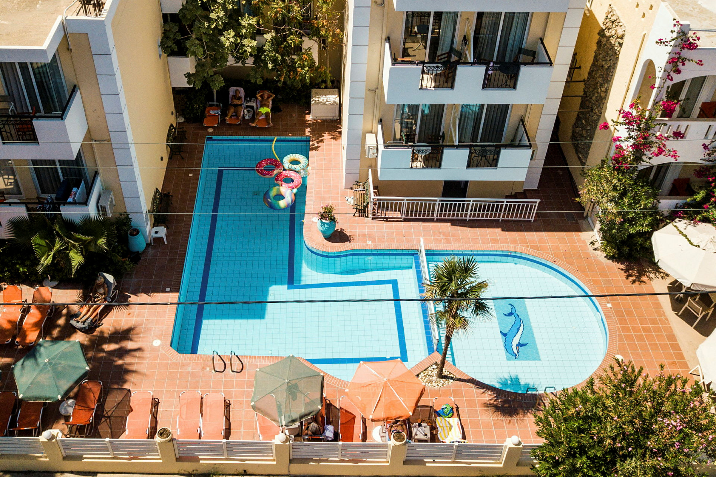Et billede af en swimmingpool med liggestole og parasoller, omgivet af hotelbygninger. (AI-genereret tekst)