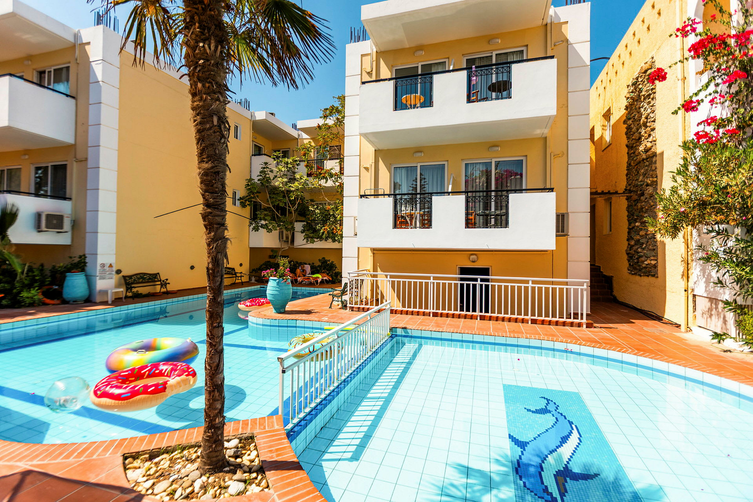 Billede af en bygning med en swimmingpool foran. Poolen har en dekoration af en delfin og flydende genstande, omgivet af en terrasse med planter og havemøbler. (AI-genereret tekst)