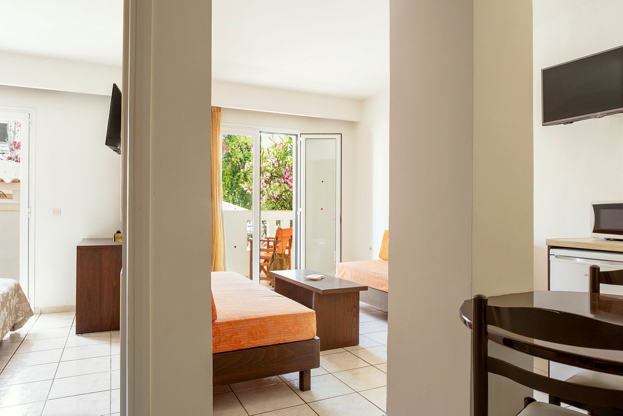 Et hotelværelse med åben planløsning, møbleret med orange puder, bord og stole samt et fjernsyn. Døren til en balkon med udsigt til grøntområder ses i baggrunden. (AI-genereret tekst)